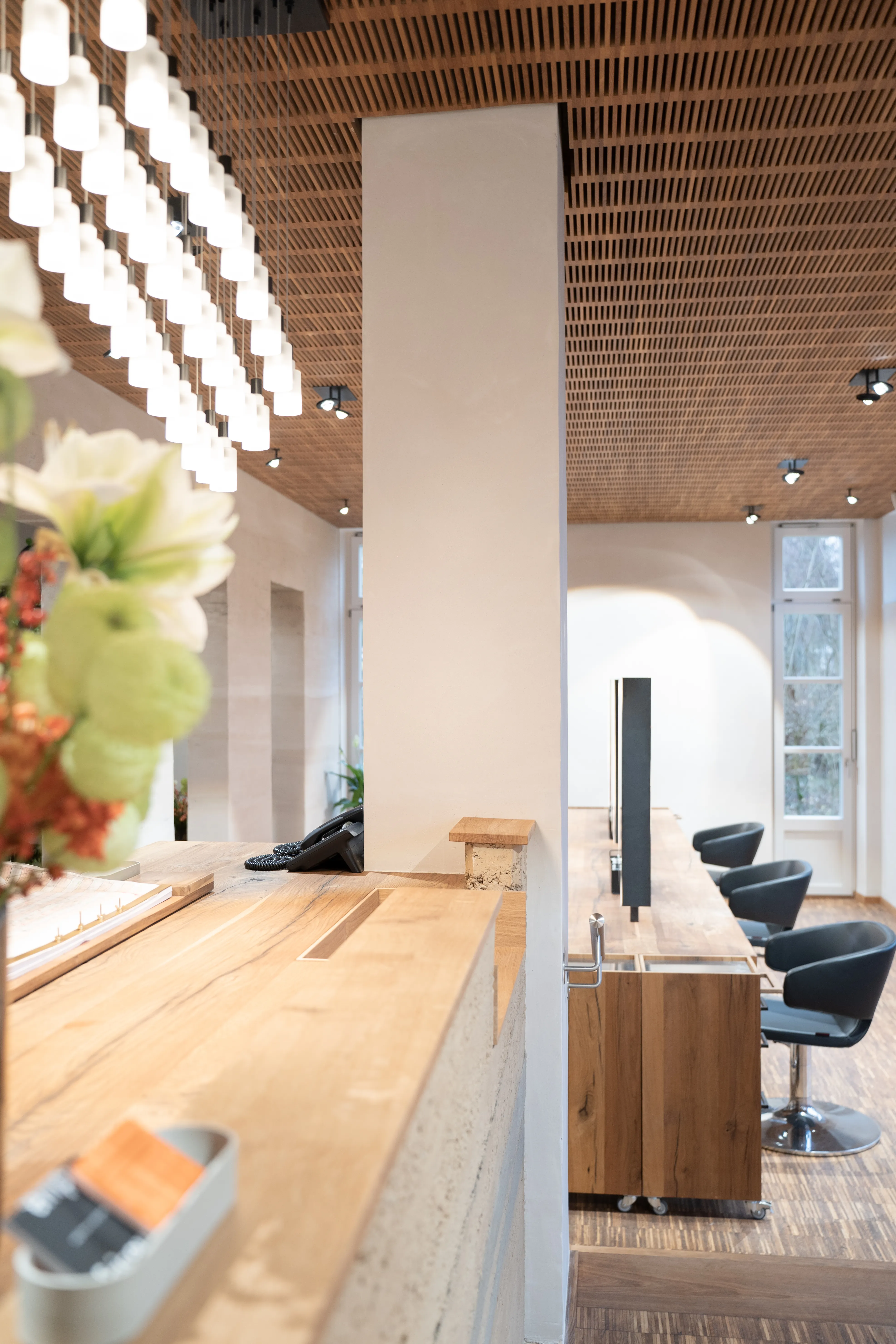 Styling area at EMPL FRISEURE with black salon chairs at an oak counter, a grid of frosted glass pendant lights and a wooden slatted ceiling.