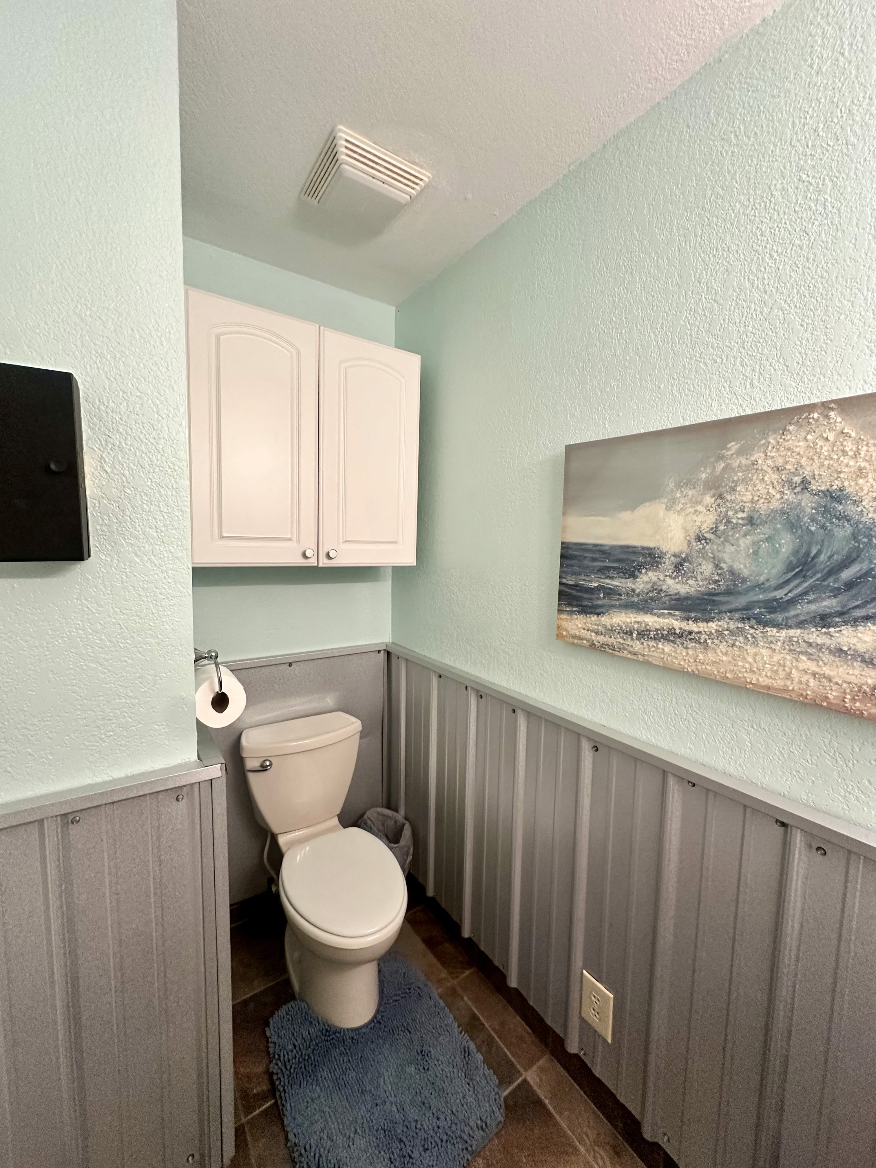 Small bathroom with light blue walls, featuring a white toilet, a toilet paper holder, a textured gray rug, wall-mounted white cabinets, and a wave-themed ocean artwork above wainscoting.