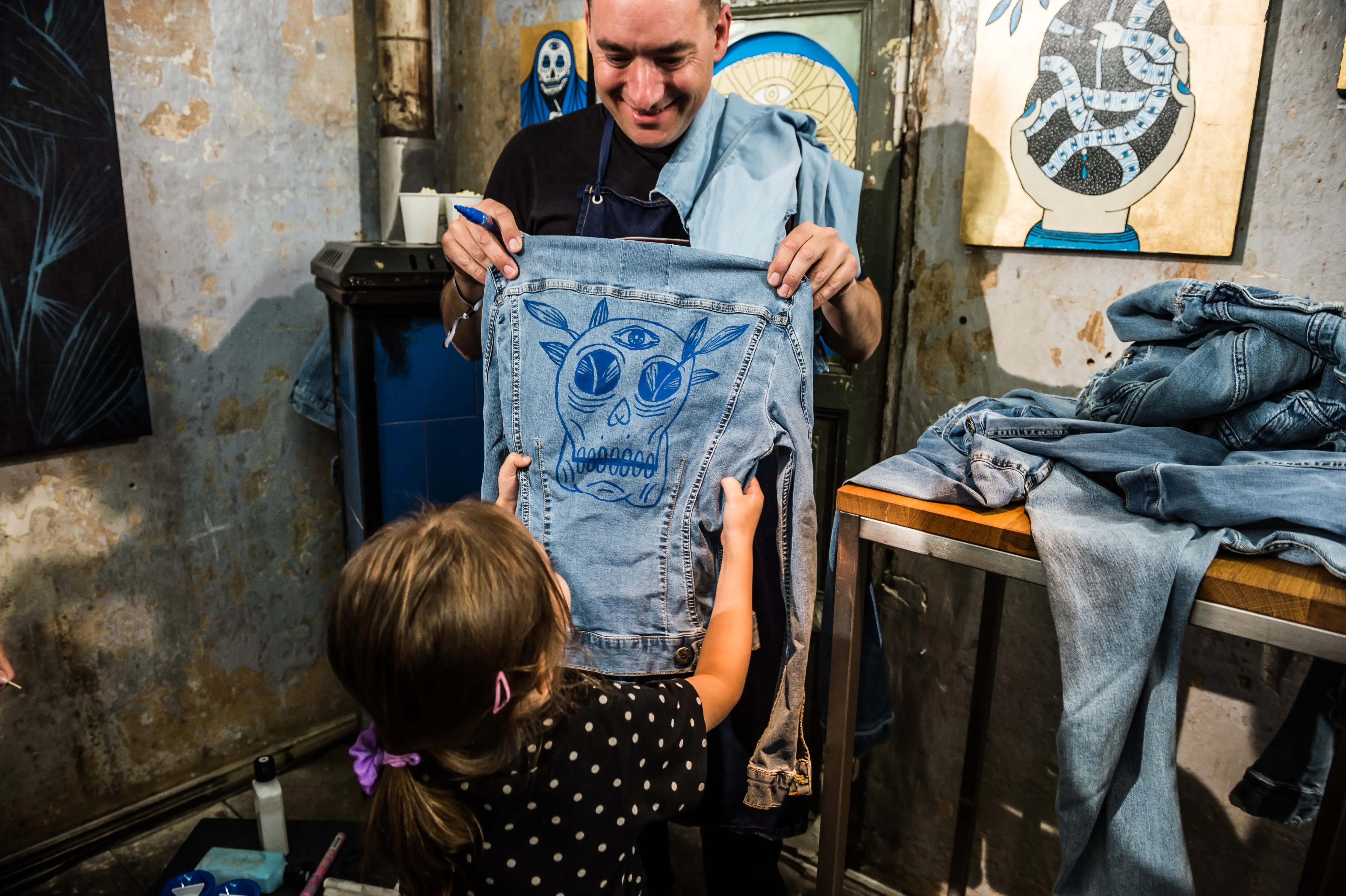 Artist showing a child a denim jacket painted with a blue skull motif at the MUSTANG Street ART Lab workshop.