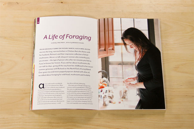 An open book lies on a light wooden surface, displaying text on the left page titled "A Life of Foraging" and a photograph of a woman in a black top preparing food in a kitchen on the right page. The woman is standing at a counter with kitchen items visible. The book is positioned at a slight angle, revealing both text and image content.