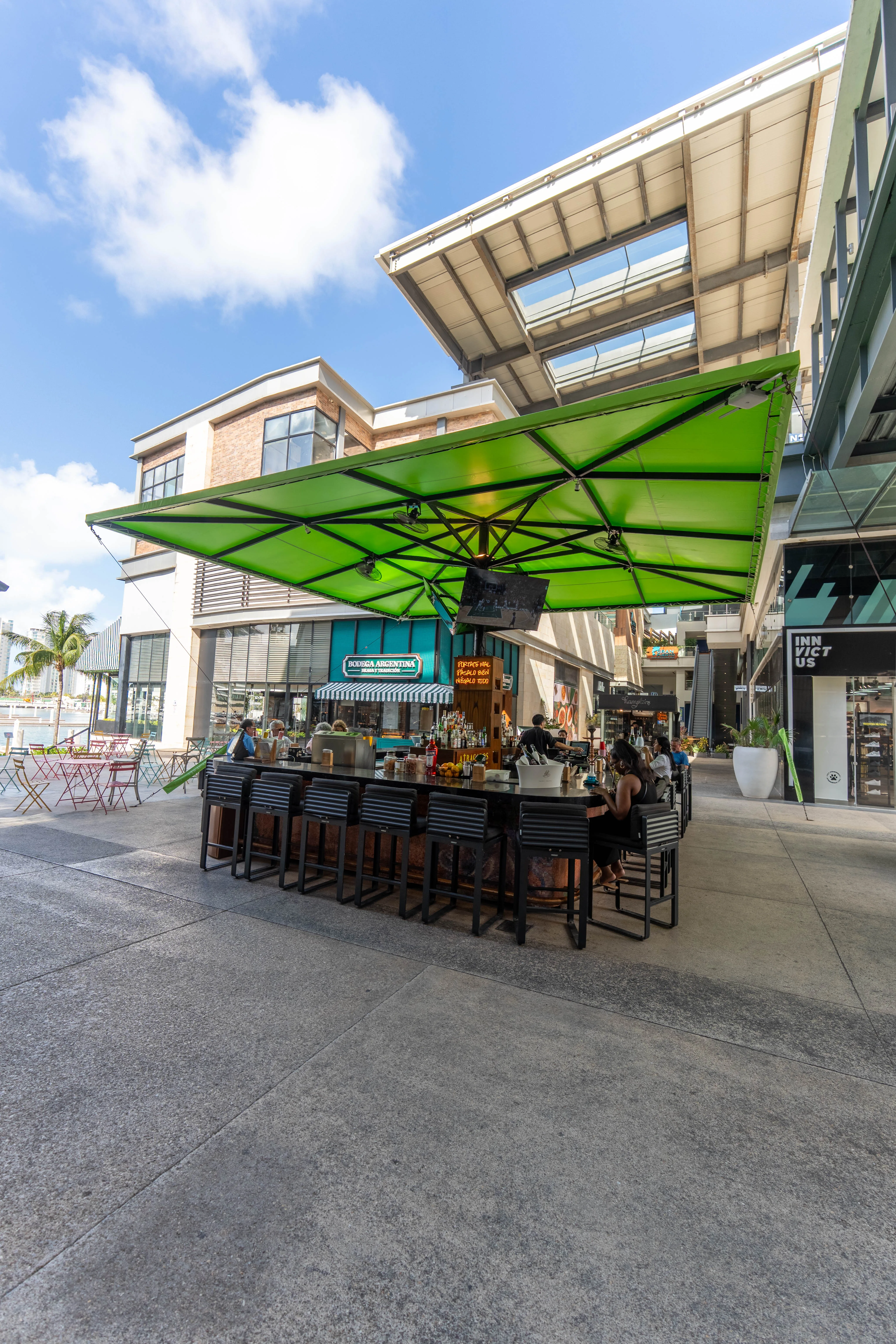Terraza exterior de Atracados con sombrillas verdes, mesas de bar, clientes disfrutando, arquitectura moderna con techo de cristal y palmeras