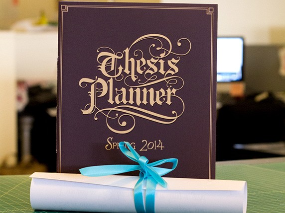 A photo of a book cover with title Thesis Planner made from stylish medieval fonts. Near the cover, placed also on the green table, there is a scroll pf paper tied with a cyan ribbon.