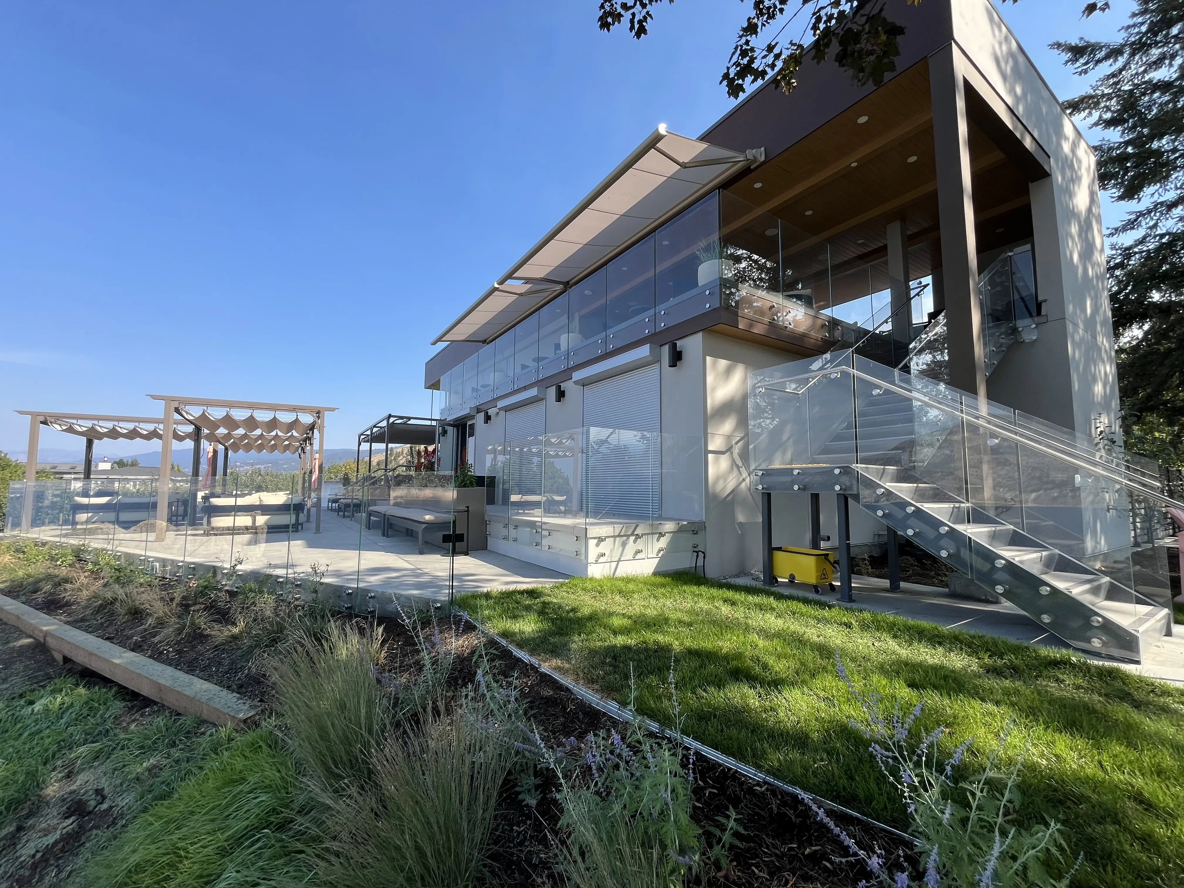 A contemporary multi-story hospitality building with extensive outdoor patio spaces. The upper balcony is lined with a seamless glass railing system under a canvas awning, while a structural outdoor metal staircase features heavy-duty glass guardrails secured with stainless steel standoff pins next to a landscaped lawn.