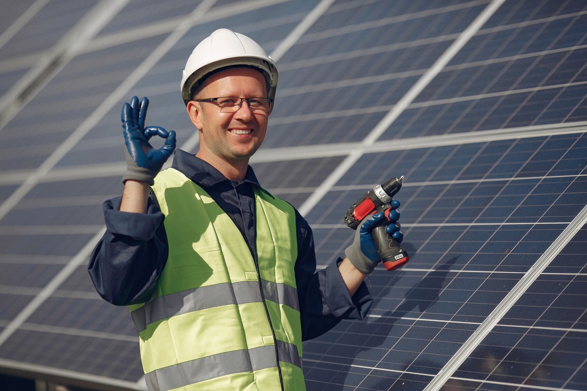 A worker in a hard hat and safety vest holds tools, smiling in front of solar panels.