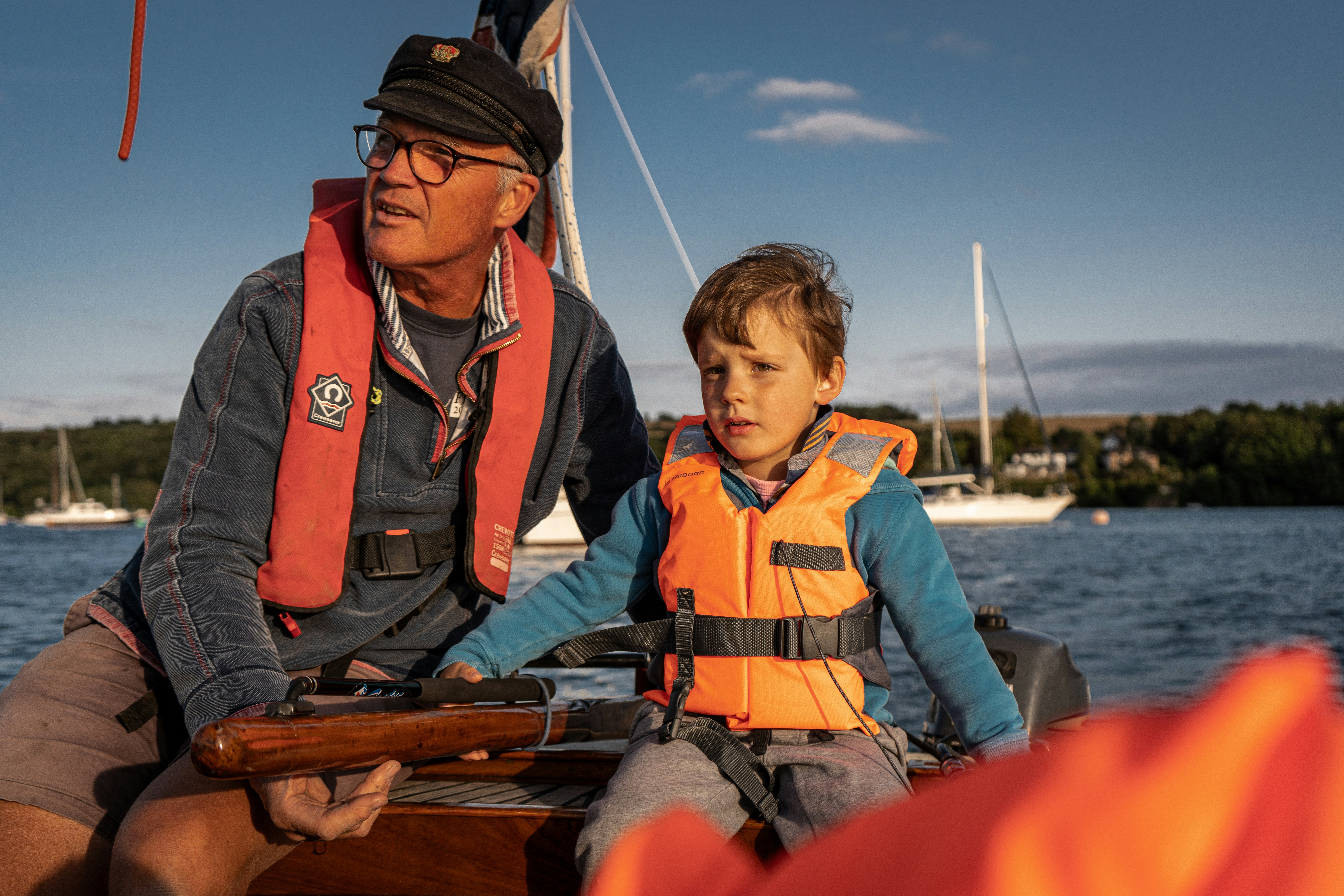 An older man and young boy, both wearing life jackets, enjoy a sunny day sailing on a tranquil lake with sailboats and a green shoreline in the background.