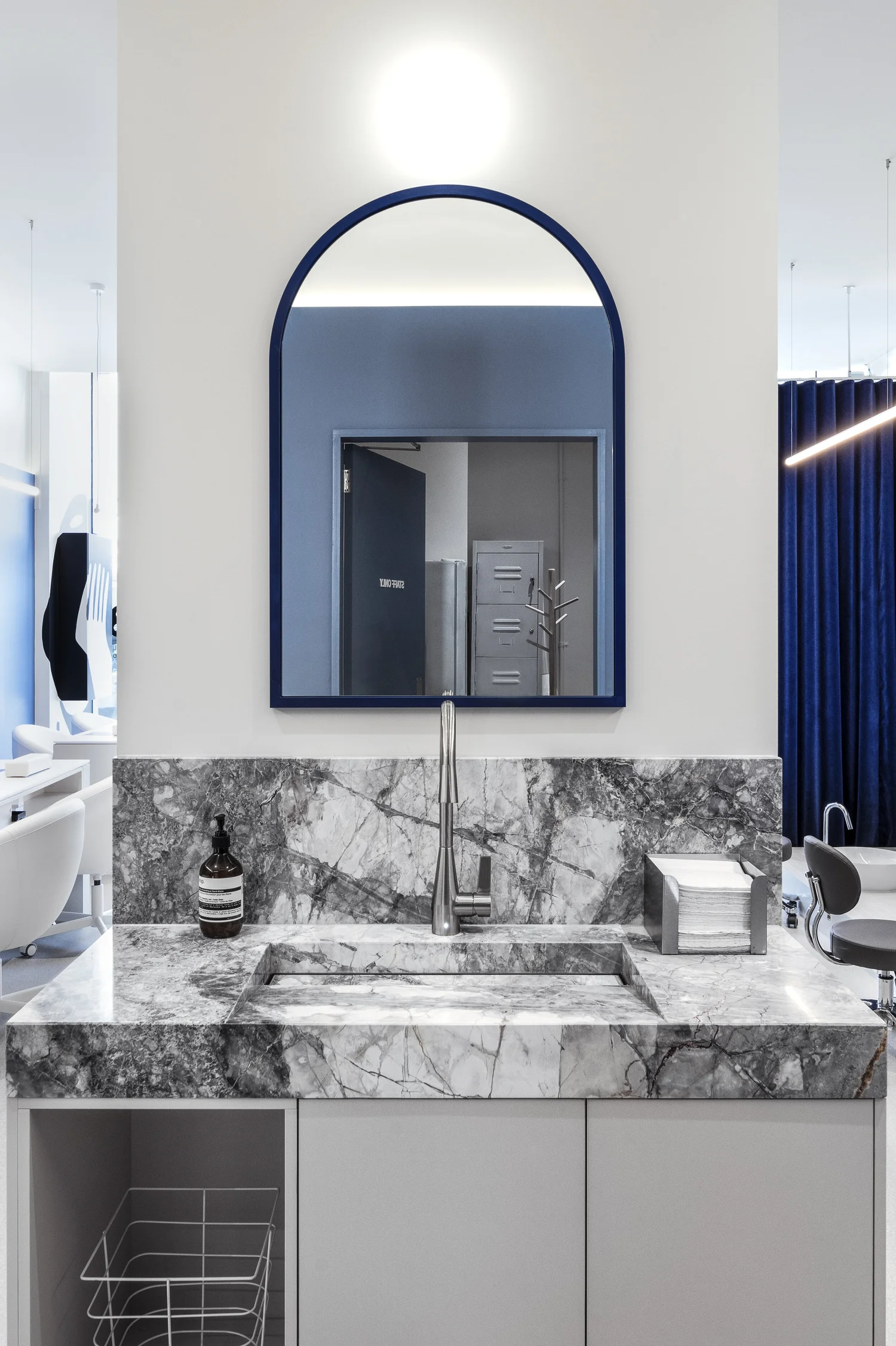 Wall-mounted arched blue mirror above a marble vanity with a soap dispenser, tap and folded hand towels, reflecting a staff-only storage room