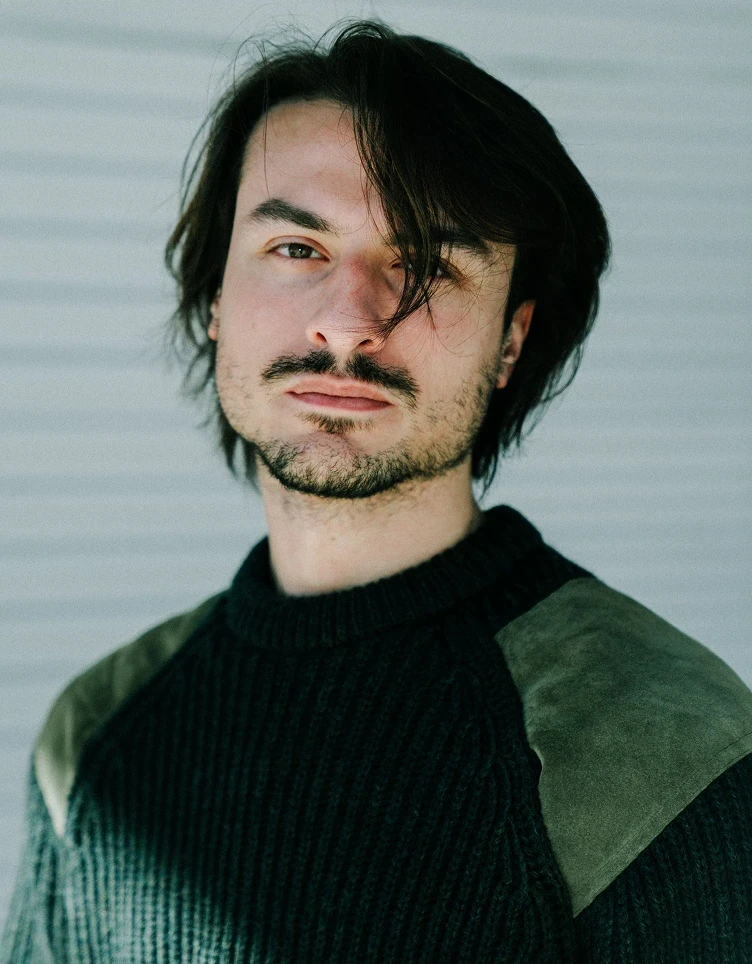 A contemplative man with medium-length brown hair and a beard wears a dark green knit sweater. He gazes seriously into the camera in soft daylight.