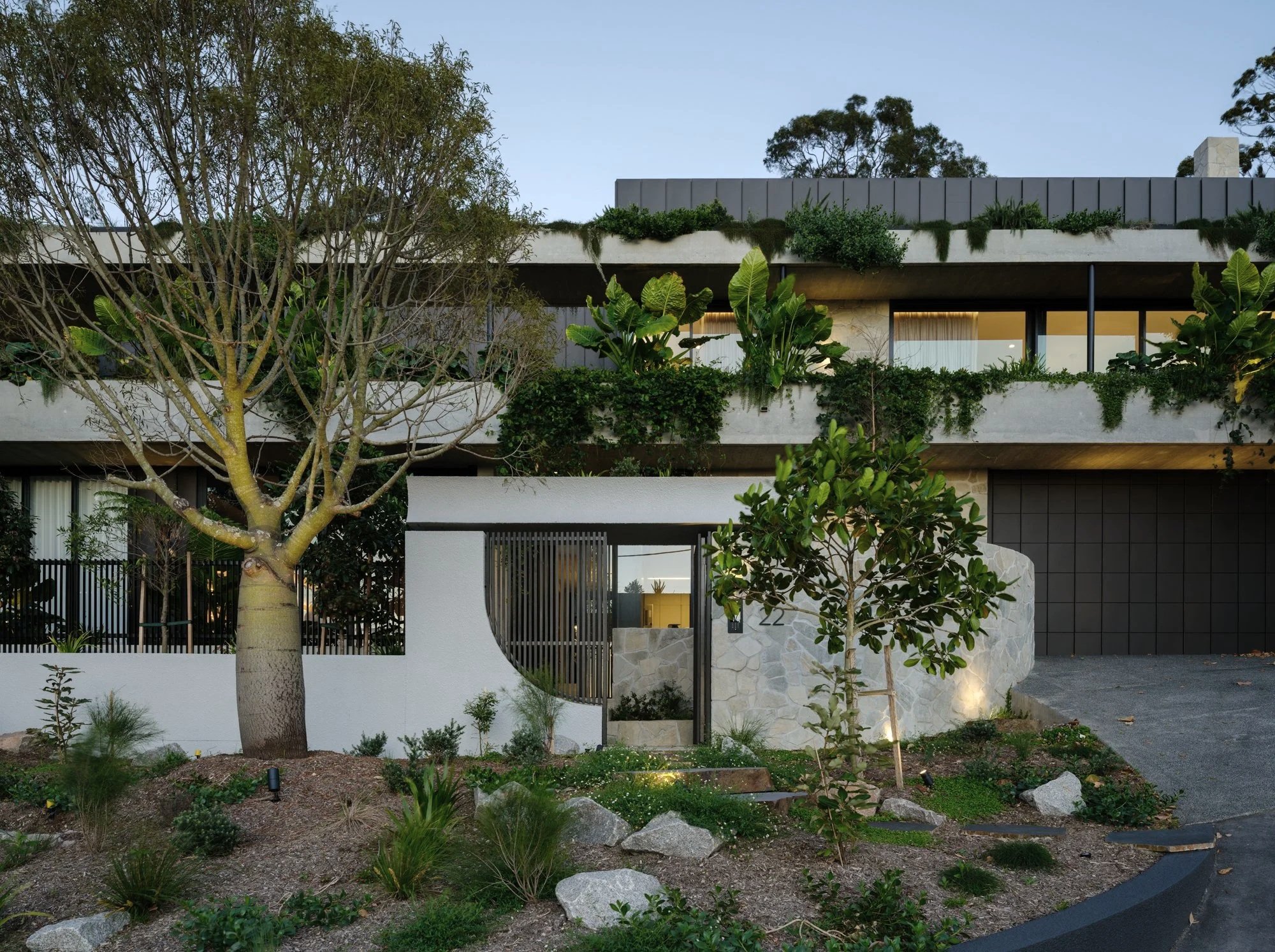 Layered concrete façade with integrated greenery, curved entry wall, and gated front courtyard at dusk.