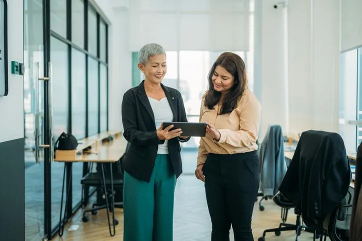 Two women in business attire looking at a tablet in a modern office setting, discussing financial strategies for debt relief and wealth building.