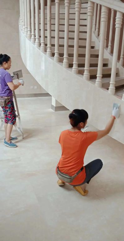 Two women cleaning the polished marble surfaces of a grand staircase in a luxury home. The staircase’s marble balustrade and flooring reflect the opulent design.