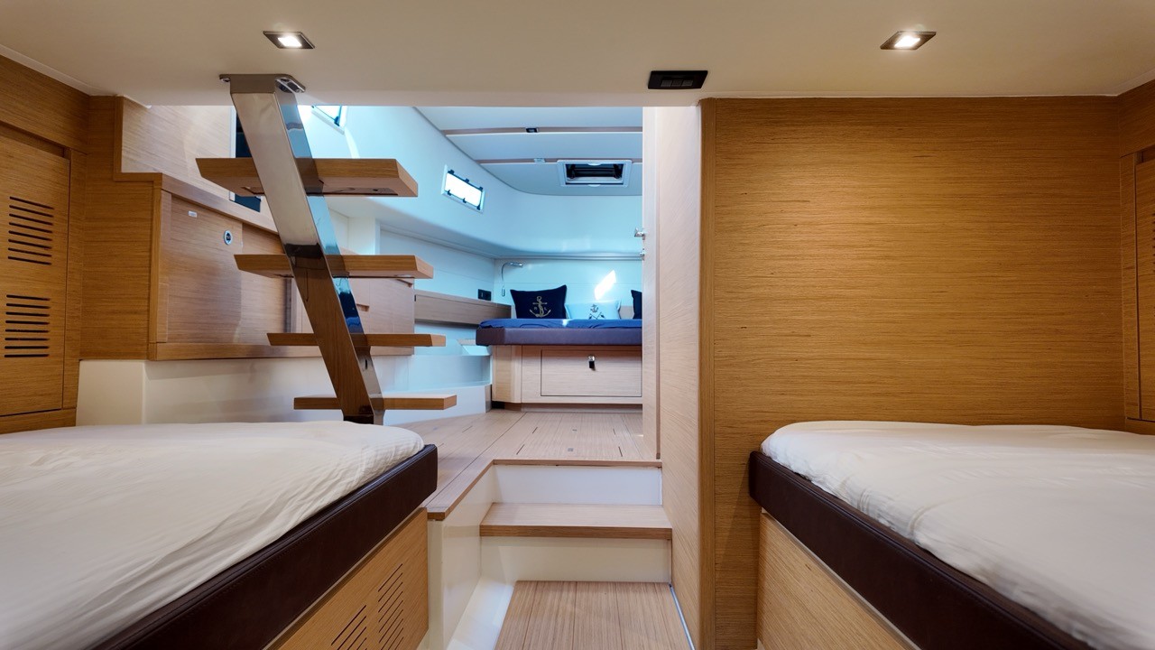Pardo 43 motor yacht forward twin cabin interior showing two single berths with white fitted mattresses, light oak veneer cabinetry with under-berth storage, floating open-tread companionway staircase in oak and stainless steel, and the forward master cabin with navy blue bedding and anchor-motif cushions visible beyond