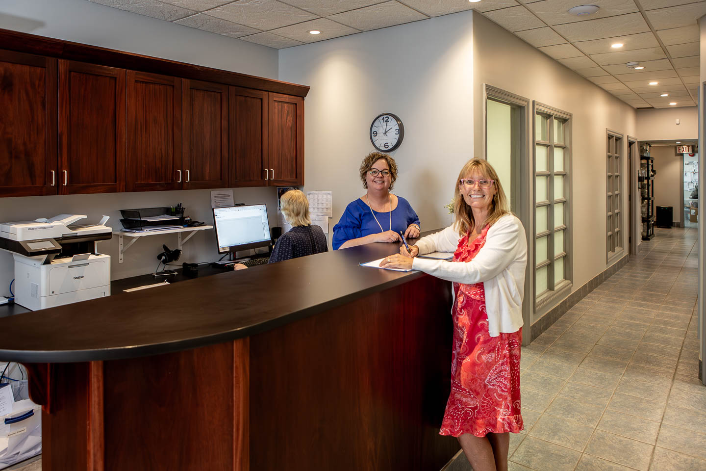 Two women on either side of a wooden reception desk looking toward the camera. One has a pen in her hand and is writing whilst smiling 