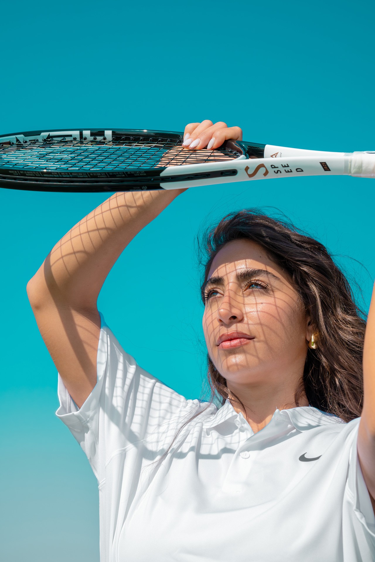 A woman in tennis whites raising a racket overhead against a clear blue sky.