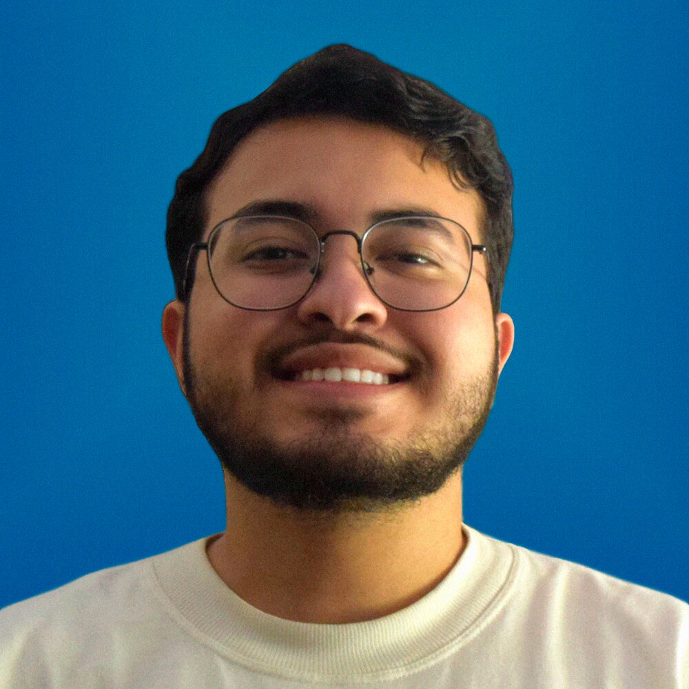 Homem branco de óculos redondos, barba pequena e camiseta branca. Ele tem os olhos e cabelos castanho-escuros e olha para a câmera com um sorriso tímido. O fundo da foto é um cinza claro e ele está vestindo uma camiseta branca.