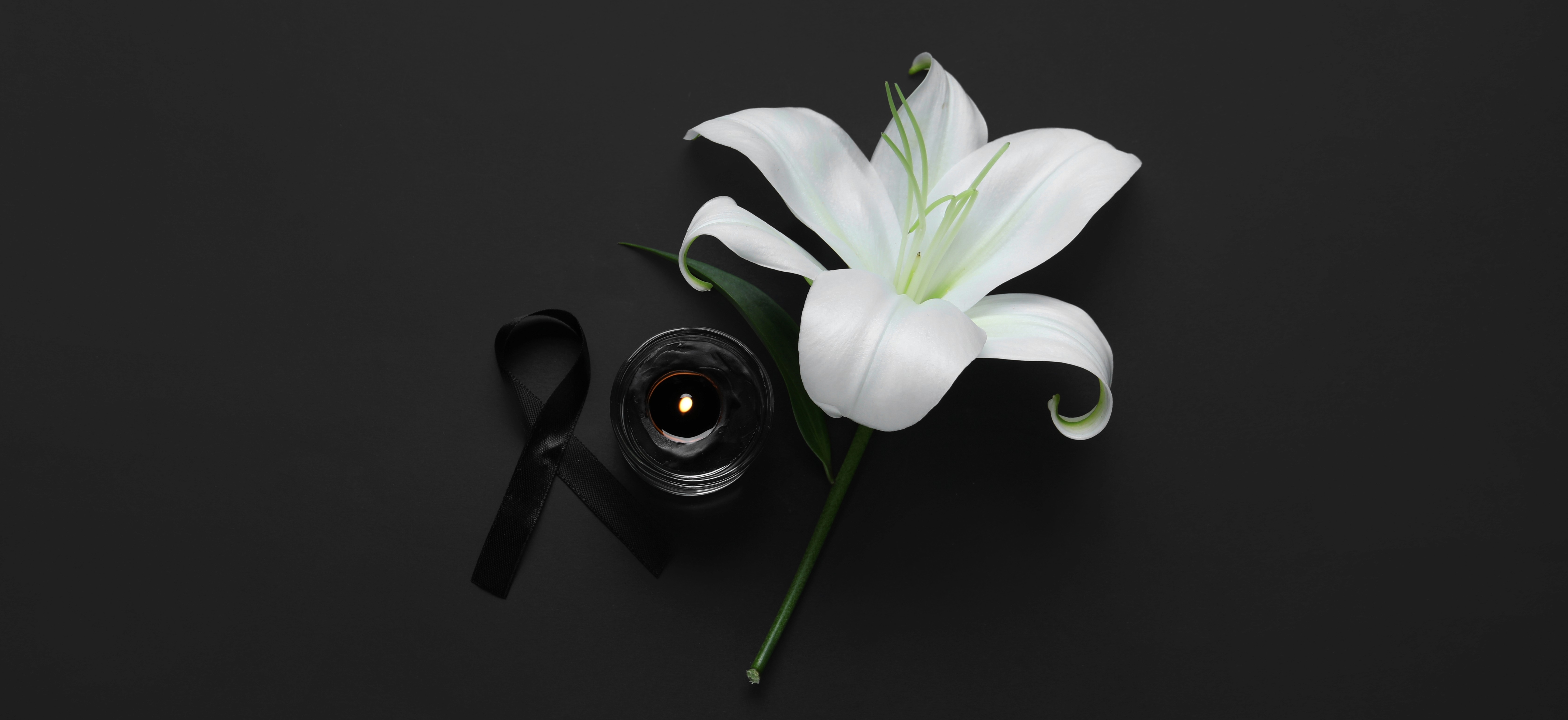 Stillleben auf schwarzem Hintergrund: Eine weiße Lilie mit grünem Stiel, eine brennende schwarze Kerze im Glas und eine schwarze Trauerschleife nebeneinander angeordnet. Das Arrangement symbolisiert Trauer und Gedenken.