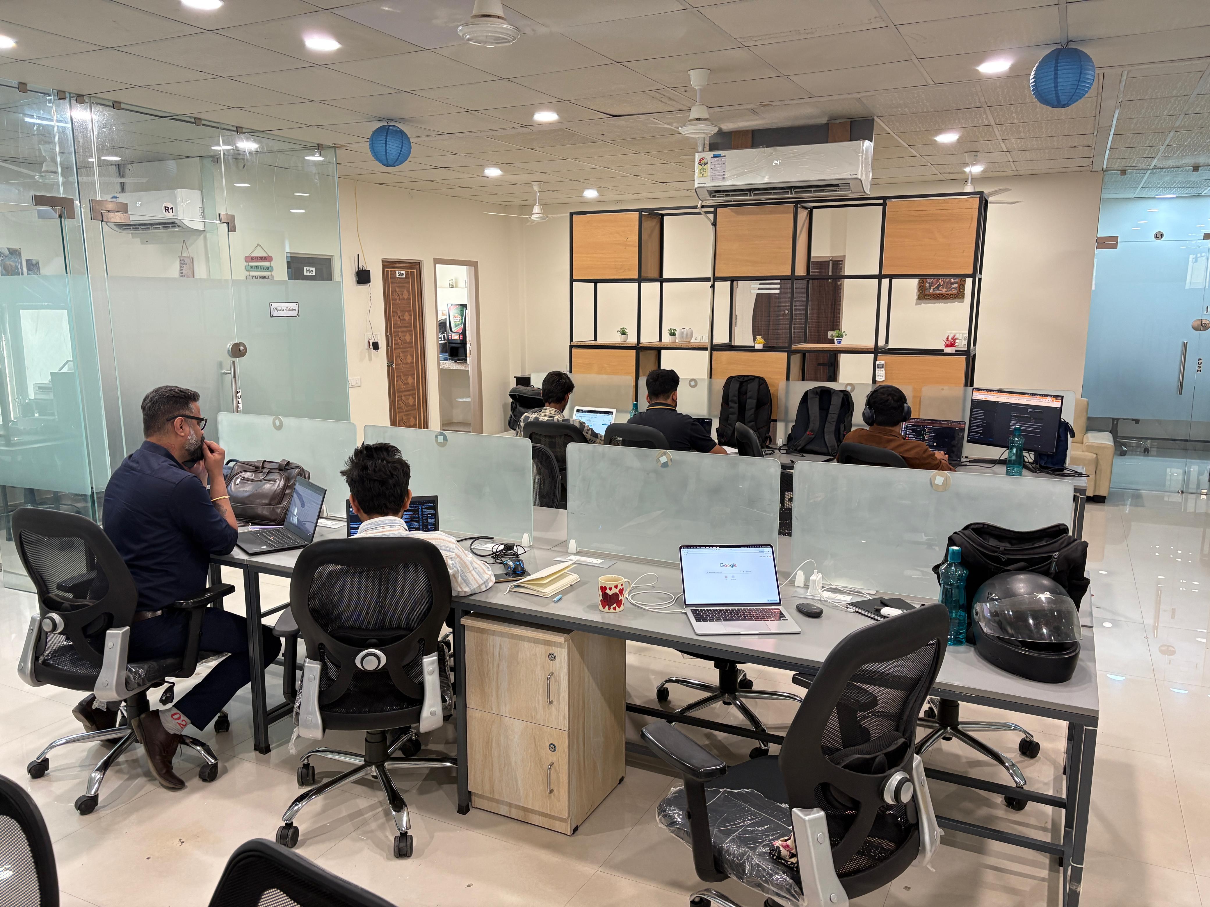 Two people having a friendly conversation at a table, one holding a mug. The atmosphere is warm with soft lighting and plants in the background.