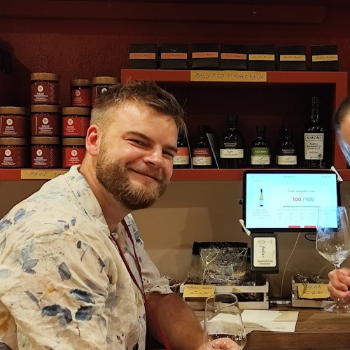 אדם בחולצת פרחים מחייך ליד דלפק בר, כשבקבוקי יין וצנצנות ברקע.