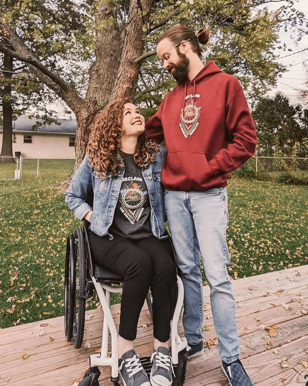 A man and woman, smiling at each other. The woman is seated in a wheelchair. They are wearing matching hoodies with a nature-inspired design.