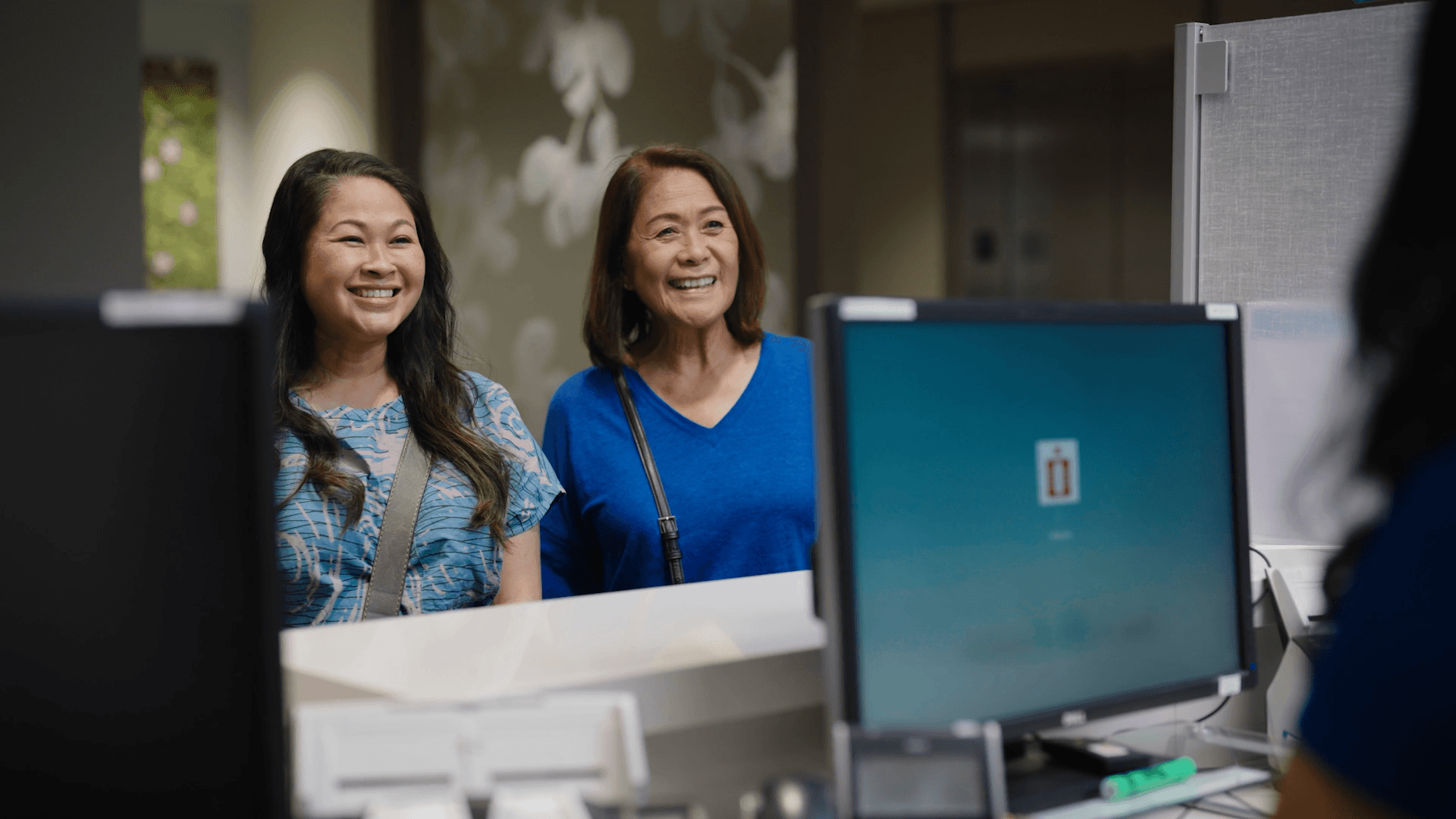 Two women greet the front desk staff inside clinic