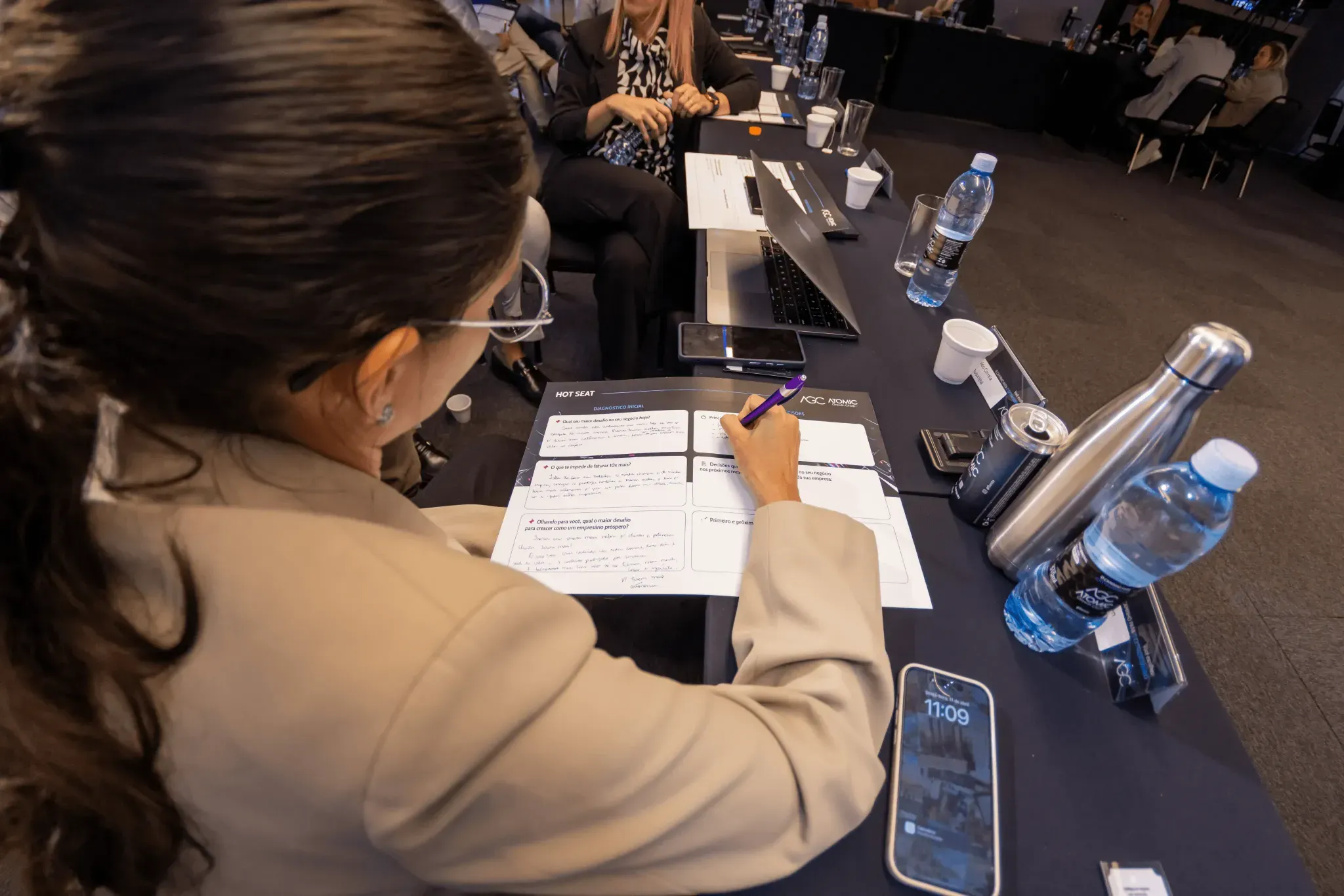A person in a beige blazer writes notes on a paper at a conference table with devices in the background. Atomic Growth Club. Atomic Group. Br24. 