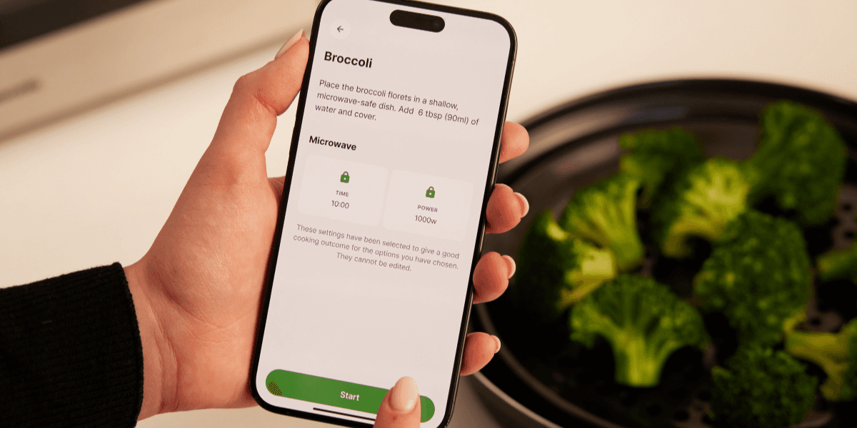 Hand holds a smartphone displaying a recipe screen titled "Broccoli" with instructions and microwave settings "Time 10:00" and "Power 1000W" beside fresh broccoli in a steamer