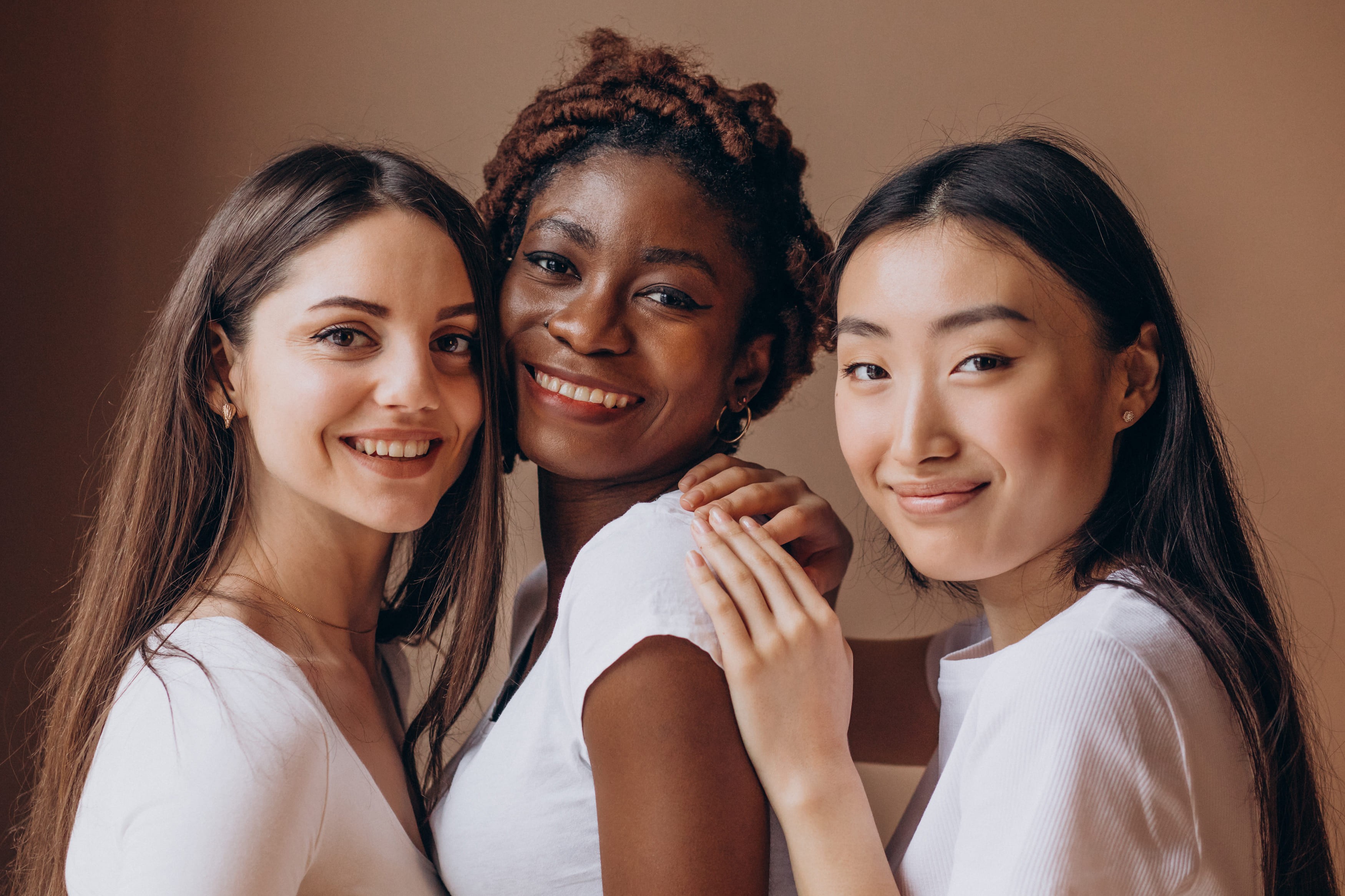 Imagem de três mulheres de diferentes origens culturais, posando juntas e sorrindo para a câmera. Elas estão lado a lado, transmitindo uma sensação de amizade e união. Todas estão vestidas com blusas claras e têm uma expressão de felicidade e proximidade, reforçando o valor da diversidade e inclusão.