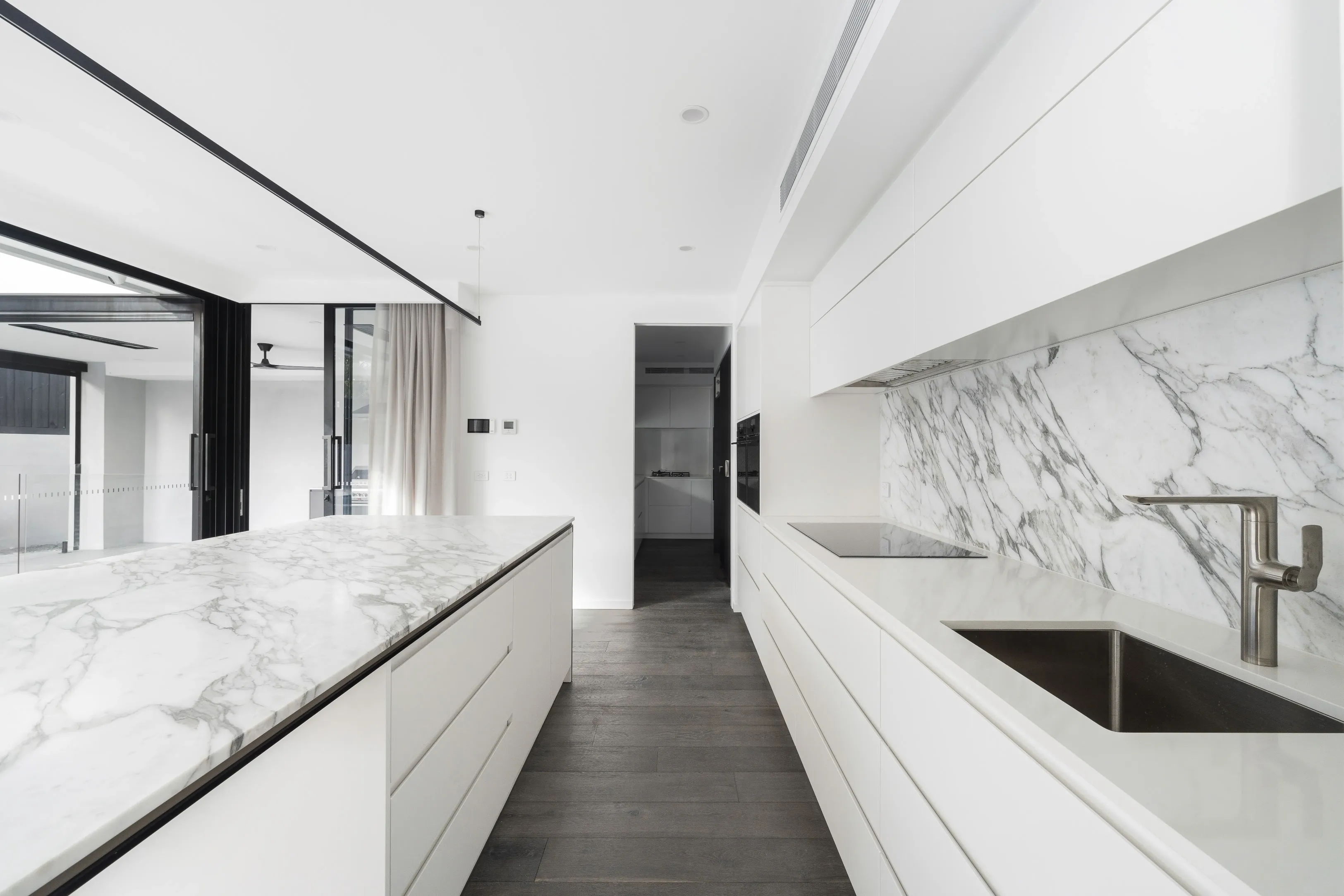 Galley kitchen with a book-matched Calacatta marble splashback, white island and cabinetry, stainless tap and sink and dark timber floors opening to the living area