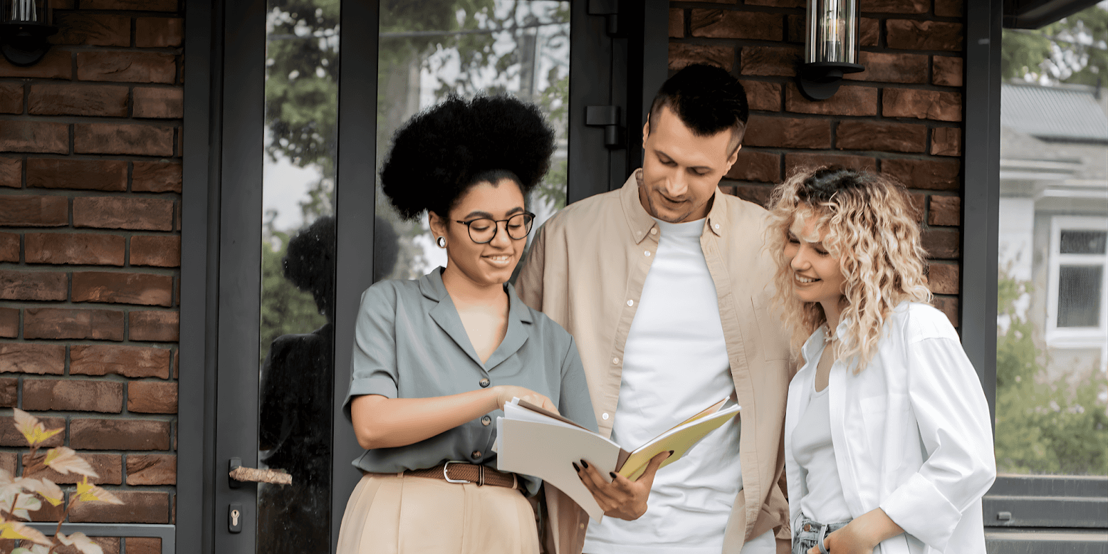 Three people stand outside a brick house, smiling and looking at an open book. The mood is friendly and collaborative, conveying a sense of teamwork.