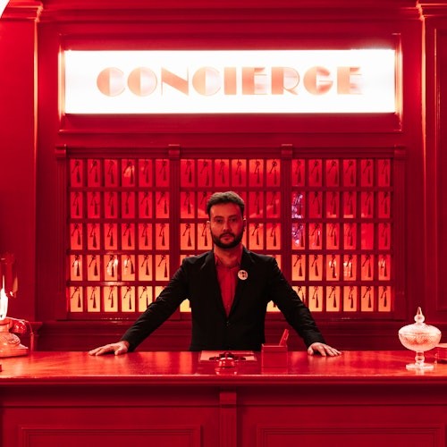 Un uomo si trova dietro un banco di concierge rosso con chiavi appese alla parete dietro di lui. Sopra la sua testa c'è un'insegna luminosa con scritto "CONCIERGE".