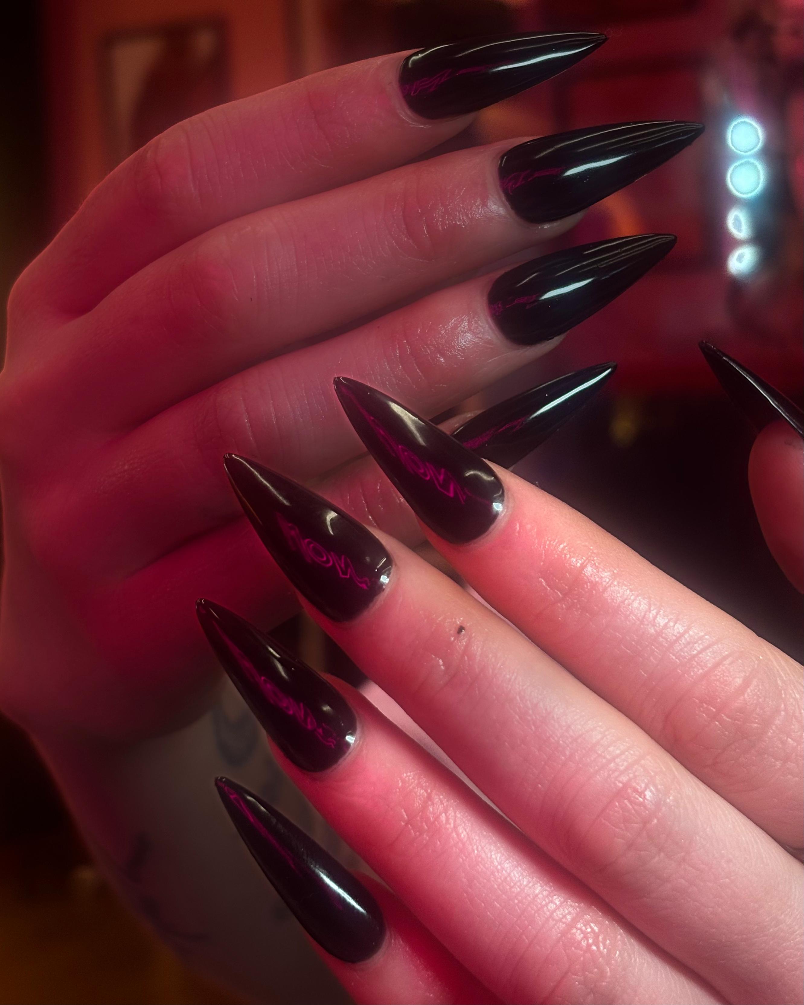 Long, sharp stiletto nails in a high-gloss black finish. Under the pink-toned lighting, subtle iridescent magenta "veins" or reflections are visible, giving them a liquid, organic look.