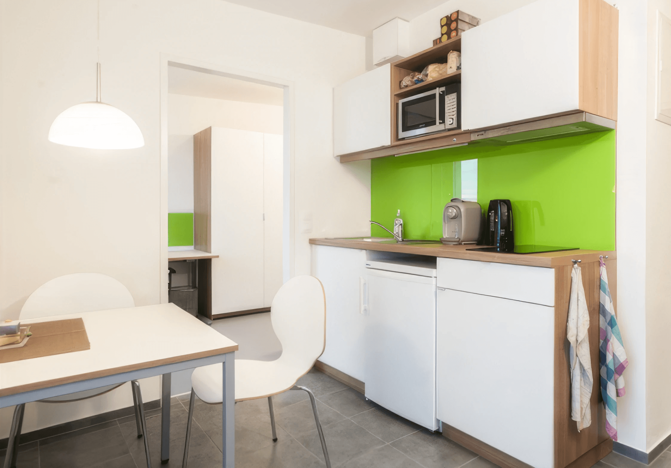 MW Interior - Kompakte weiße Einbauküche mit grüner Glasrückwand, Holzarbeitsplatte, Mikrowelle, Kühlschrank und kleinem Esstisch mit weißen Stühlen" Beschreibung: Funktionale Küchenzeile in Weiß mit leuchtendem grünen Glasrückspiegel, Holzarbeitsplatte und vollständiger Ausstattung – kompakt, klar und auf die Bedürfnisse junger Bewohner zugeschnitten.