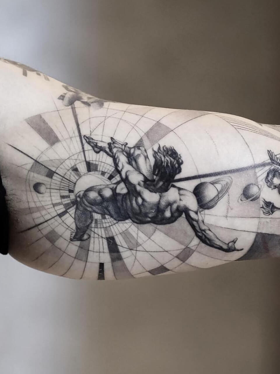 A detailed tattoo of a muscular figure with an outstretched arm, surrounded by geometric shapes and planetary motifs, covering a person's upper arm.