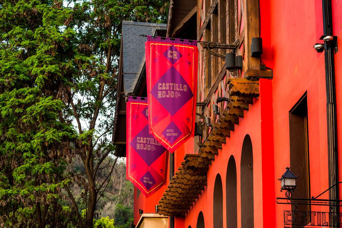 Red flags with "Castillo de Segovia" logos hang from a stone balcony of a building against a backdrop of green trees.