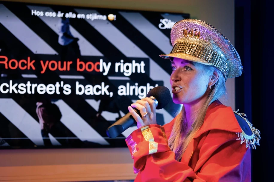 A young person with curly hair smiles at the camera while sitting in front of a laptop against a dark background.