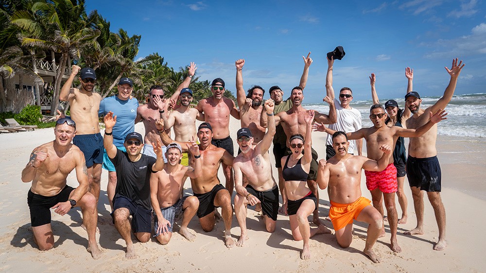 Group of people cheering on a sunny beach, with palm trees and ocean in the background.