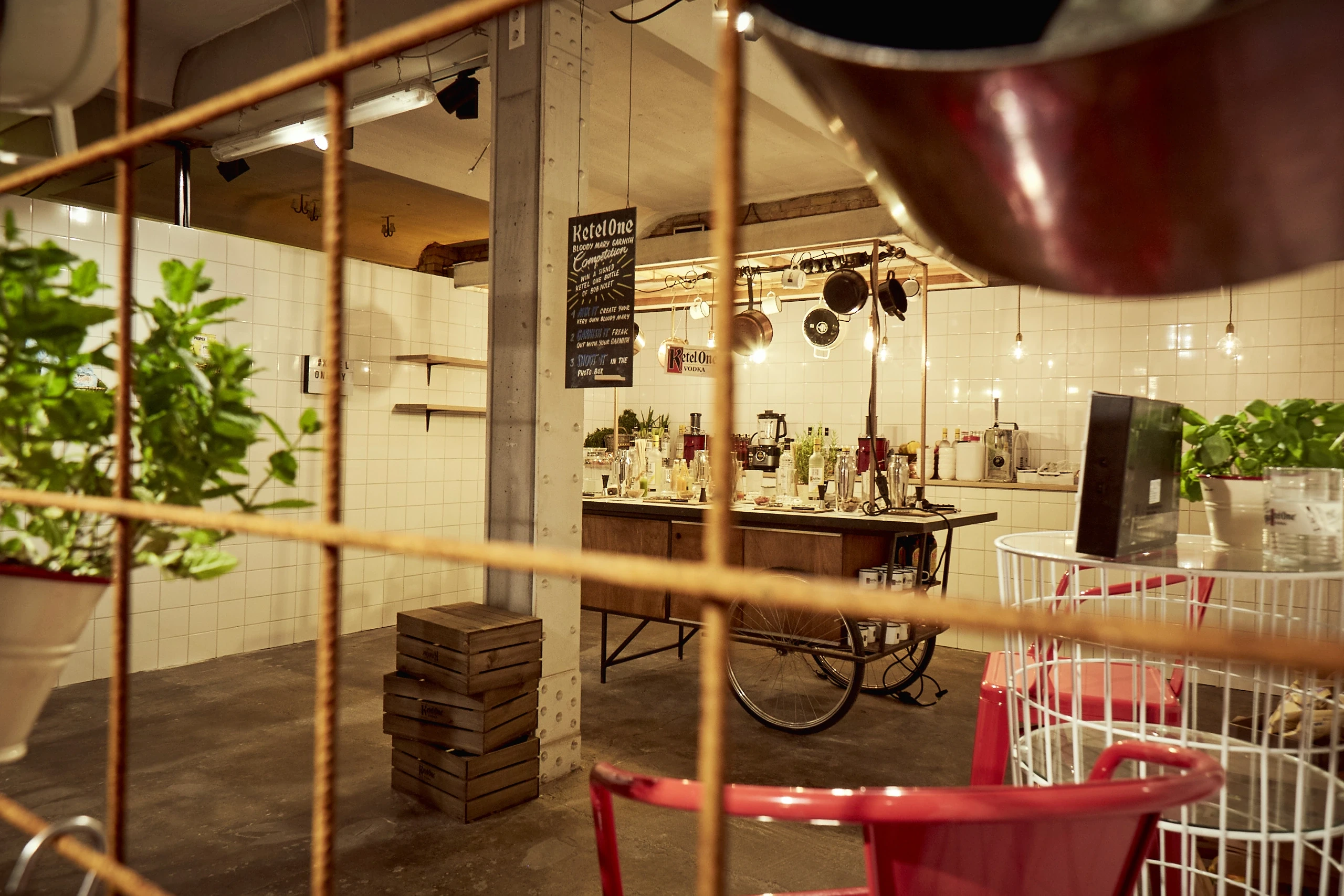View through a wire mesh into the Ketel One kitchen bar at DIAGEO BCB17 with a mobile bar cart, hanging pots, white tiles and red chairs.
