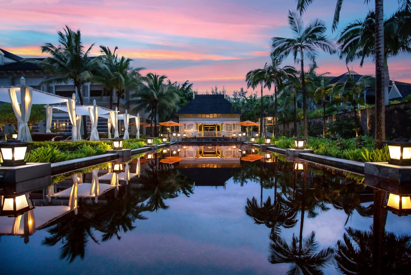 Romantische Abendstimmung an einem beleuchteten Wasserbecken im Resort, gesäumt von Palmen und Cabanas unter einem farbenfrohen Sonnenuntergangshimmel.