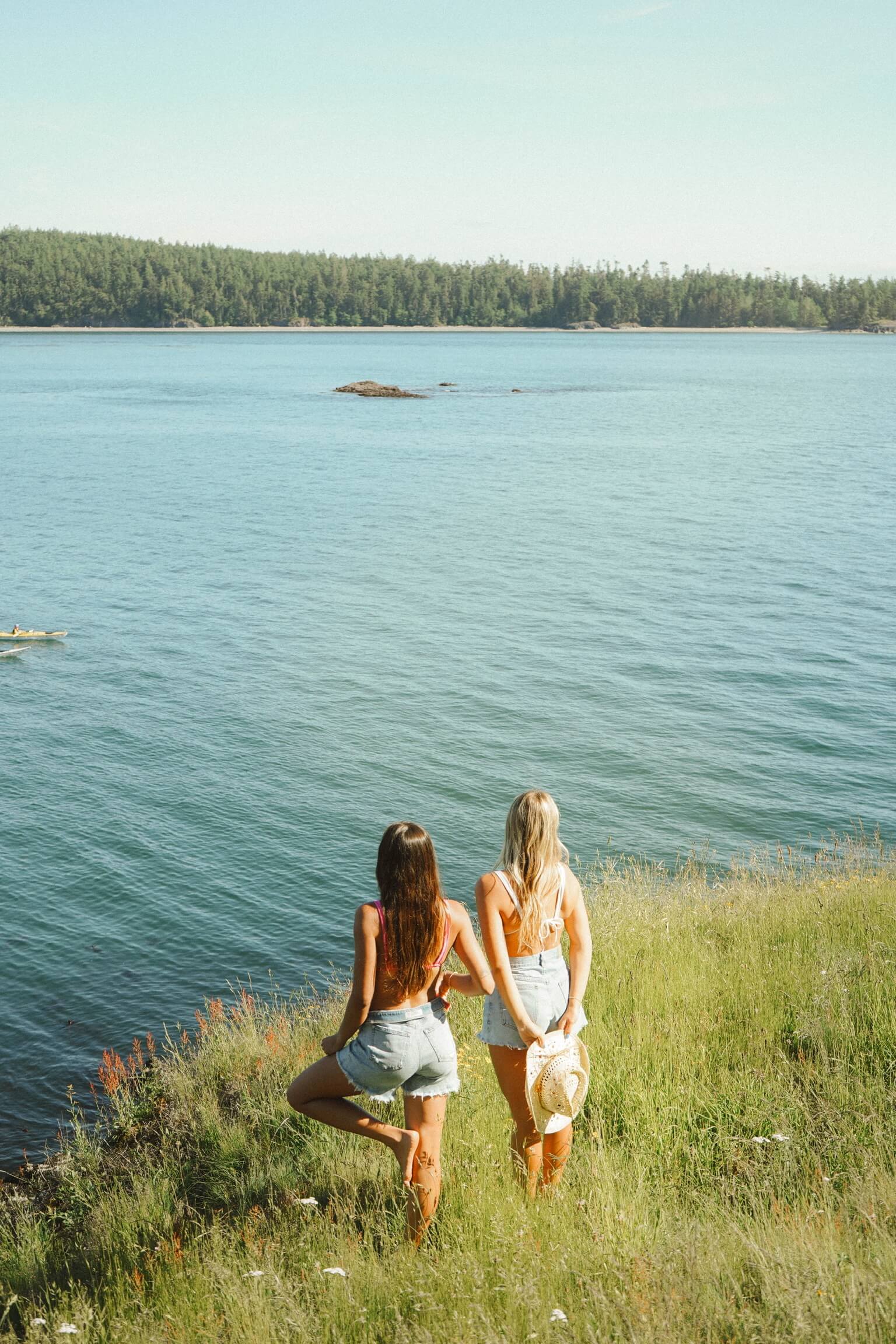 Two young women in bikini tops and denim shorts standing on a grassy meadow facing a sky blue lake. 