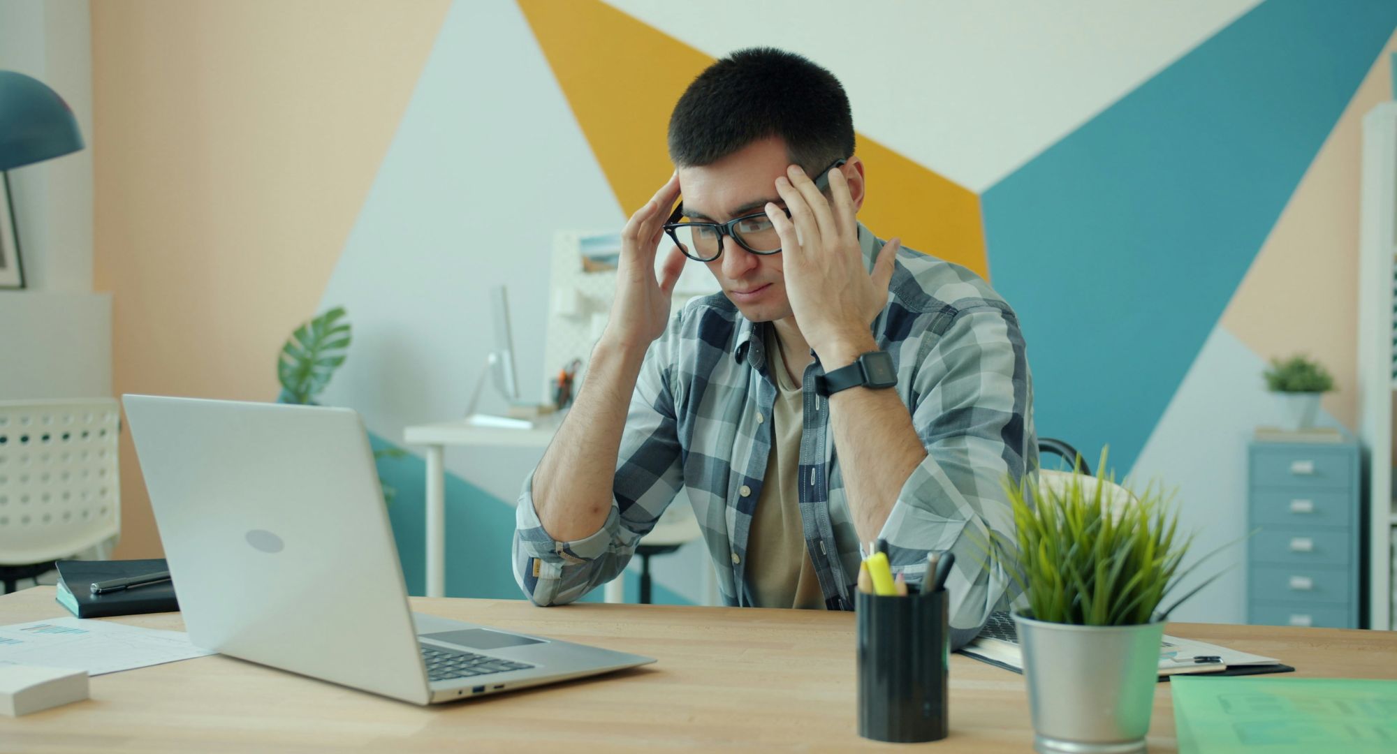 homem sentado numa mesa de escritório com notebook com as mãos na cabeça em frustação