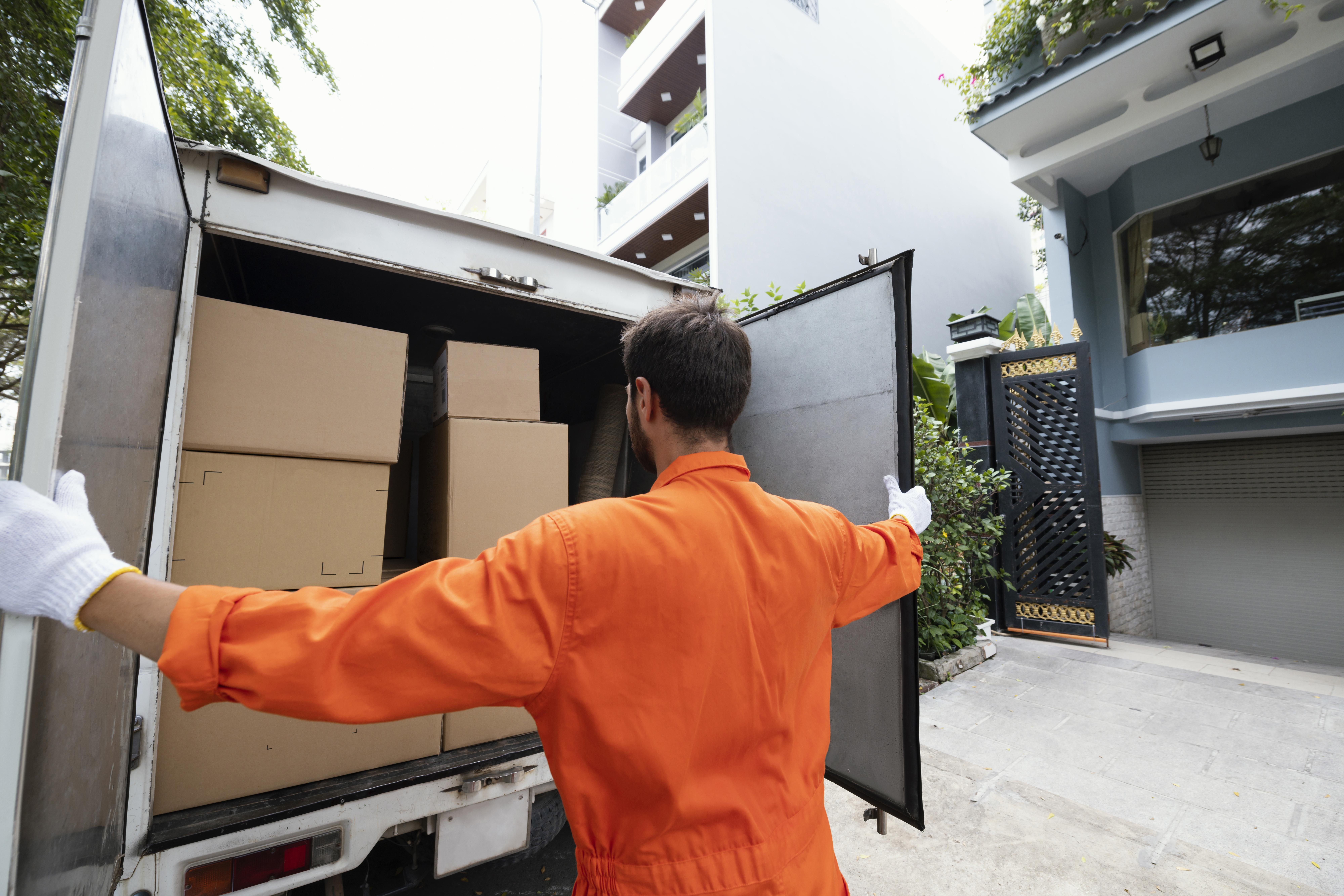 A person in an orange jumpsuit and white gloves closes a truck's doors loaded with cardboard boxes. The scene is outside a modern building.