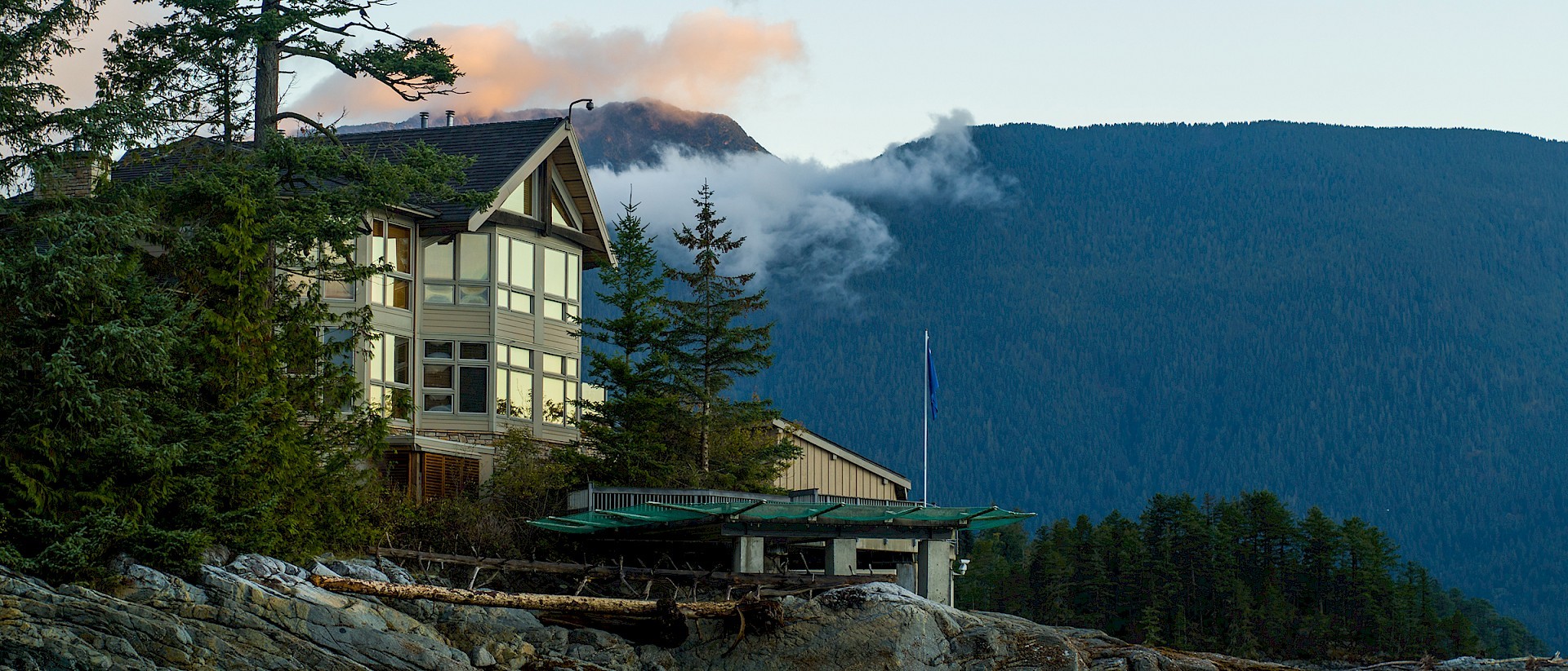 Eine rustikale Lodge aus Holz und Stein, die auf felsigem Ufer thront, umgeben von Tannen und Wolkenverhangenen Bergen im Hintergrund.
