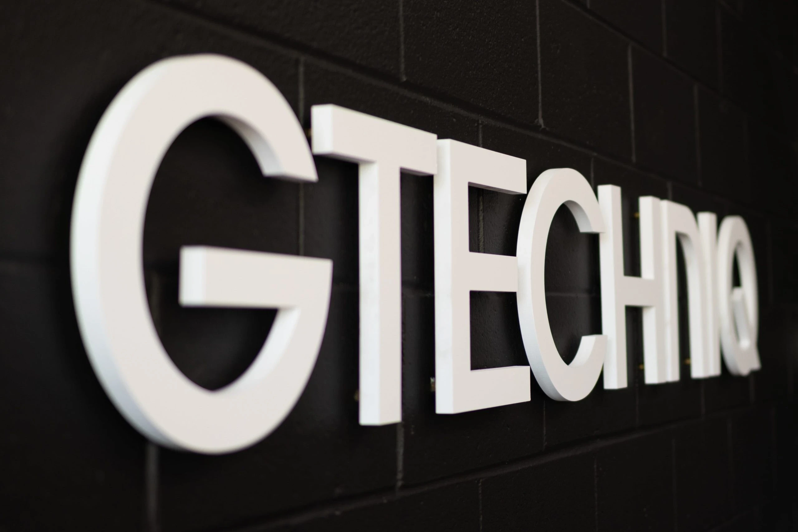 Side-angle view of the Gtechniq brand name in large white 3D letters mounted on a black wall. Sleek and professional signage for the high-tech car care company.