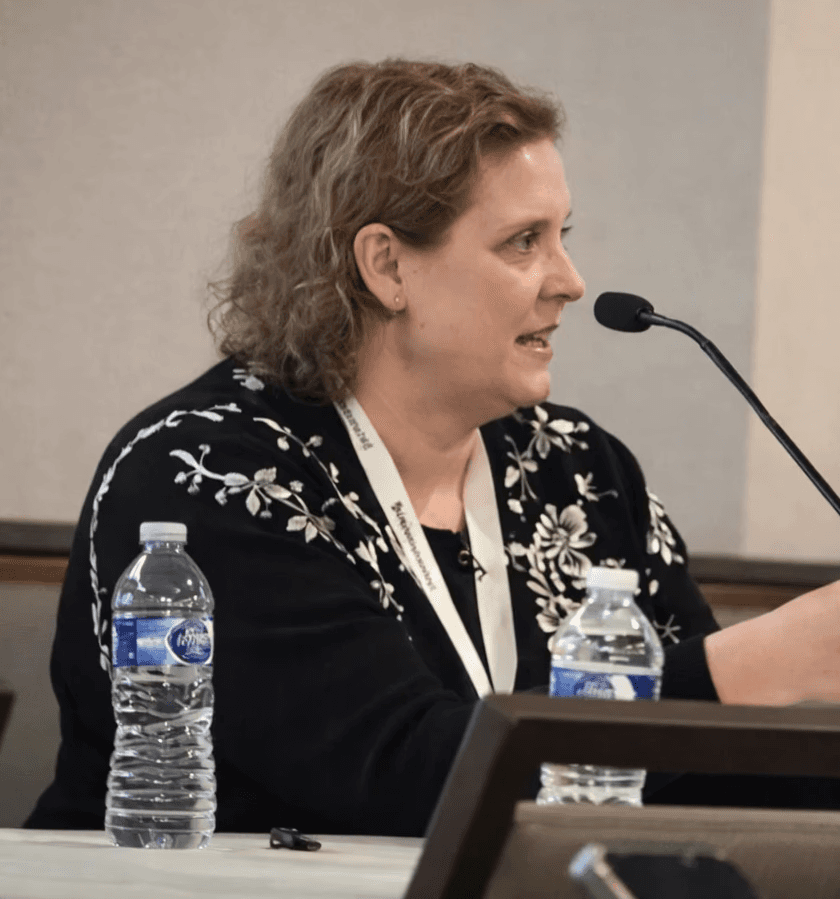 A smiling man in a dark shirt and jeans sits casually on a stool against a dark grey background, looking off to the side.