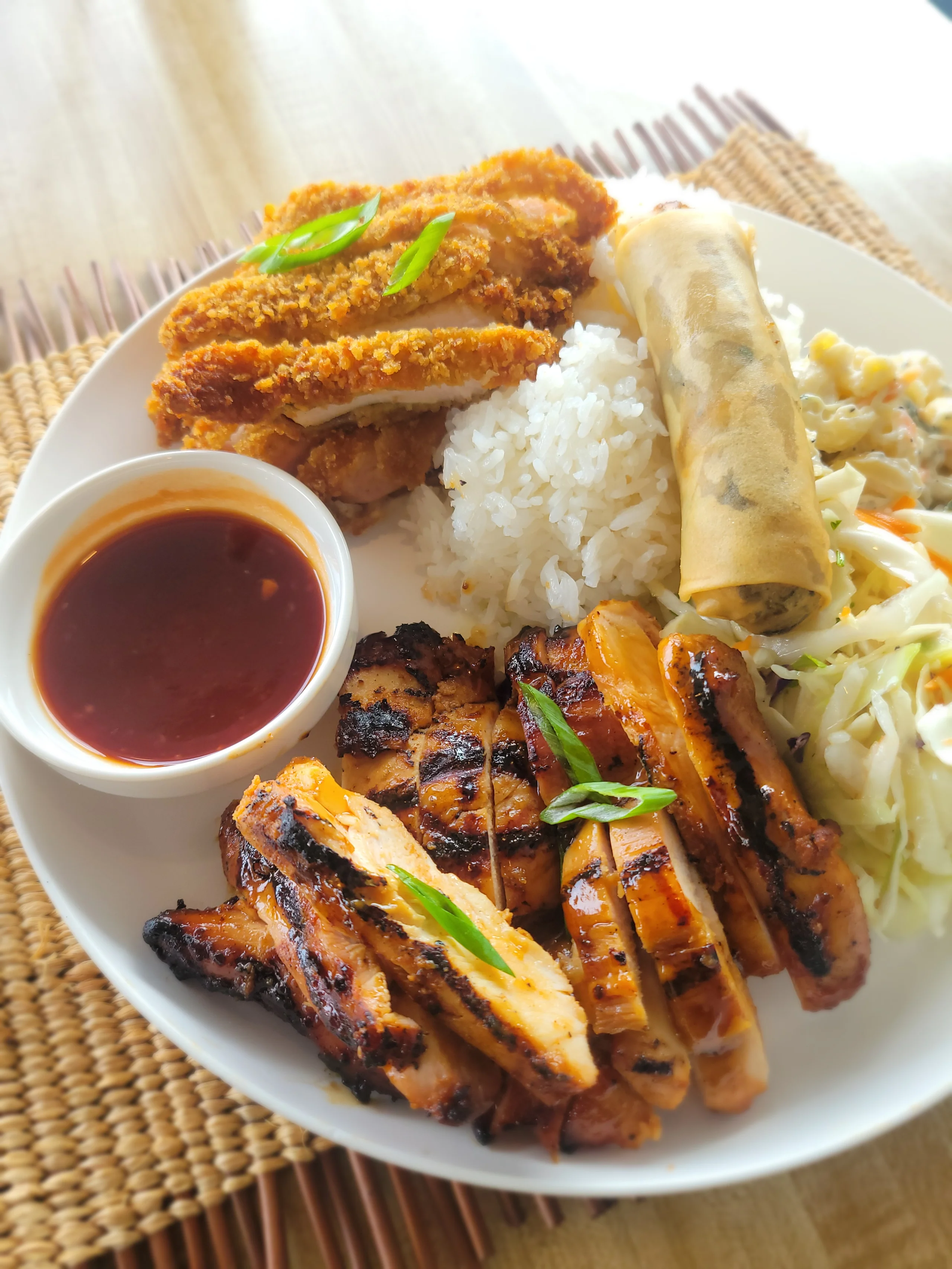 A large white plate featuring sliced grilled Huli Huli chicken and crispy golden Chicken Katsu, served with a spring roll, white rice, macaroni salad, slaw, and a side of katsu dipping sauce.