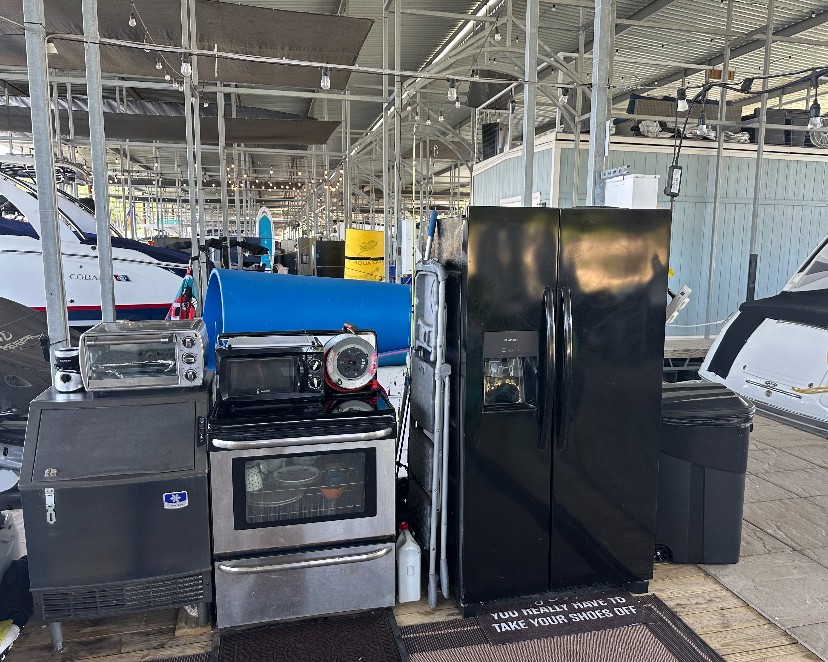 A collection of kitchen appliances, including a black refrigerator, a stainless steel oven, a toaster oven, and a microwave, is neatly arranged on an outdoor dock under a covered structure, flanked by boats on either side.