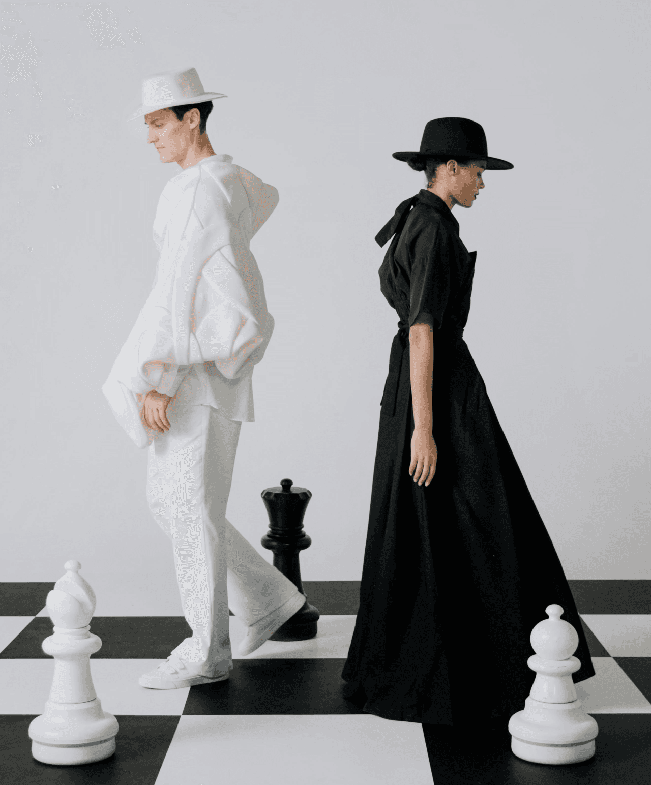 A couple dressed entirely in contrasting white and black outfits, including hats, stand back-to-back on a large checkered chess board surrounded by oversized chess pieces.