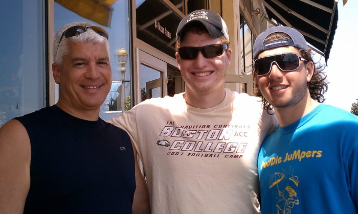 Three men smiling outdoors near a restaurant, all wearing sunglasses; the man in the middle wears a “Boston College” T-shirt, and the man on the right wears a “Double Jumpers” shirt.