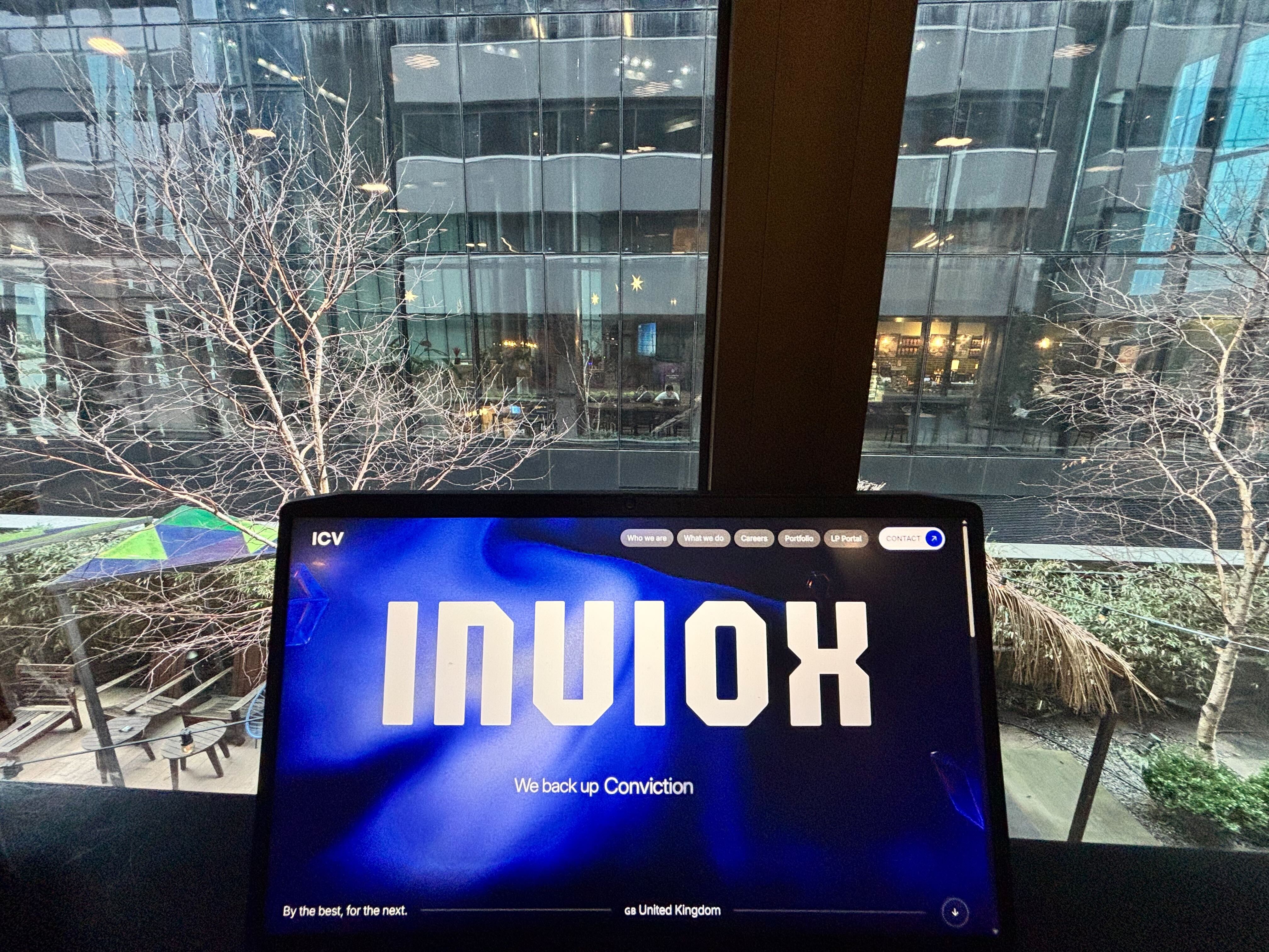 A laptop displaying an Inviox's logo, placed on a table near a window with a city view outside.