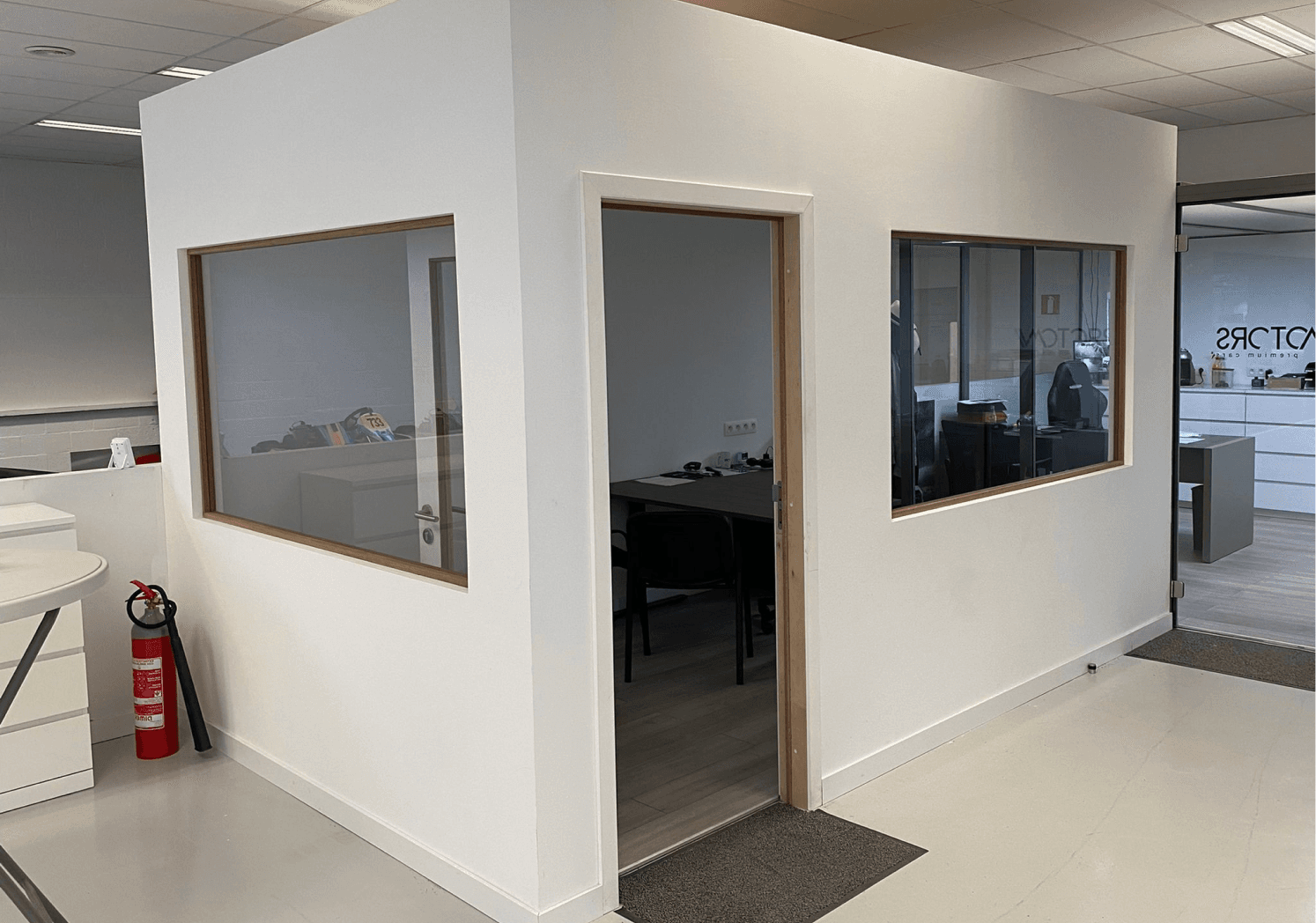 Kantoorgang met twee kamers zichtbaar door grote glazen ramen met licht houten frames, witte muren, lichte vloer en een rode brandblusser aan de linkerkant. Schone en professionele uitstraling.