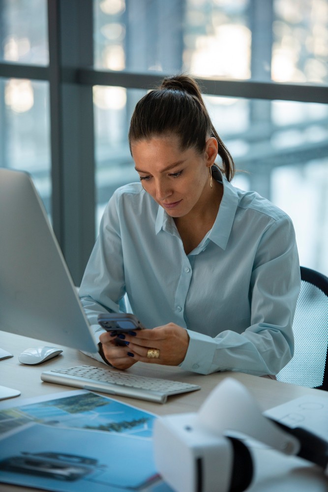 Professional woman working at a desk using a smartphone and laptop, representing connected workflows and mobile operations.